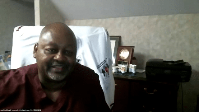 Man sitting in a hospital bed, smiling, with medical supplies and a small clock on the bedside table behind him.
