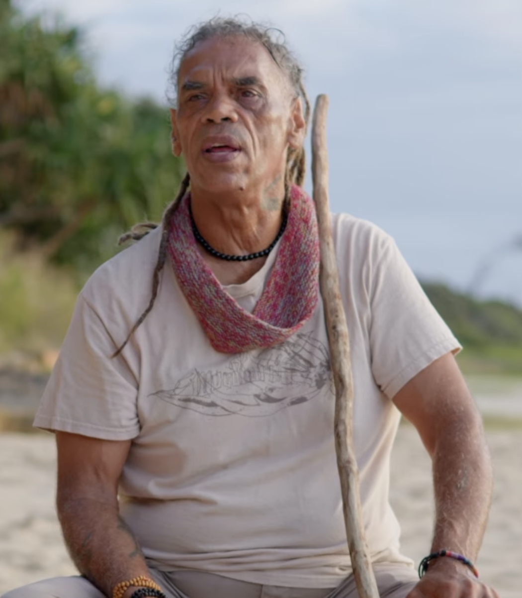 A man with dreadlocks and tattoos outdoors, holding a walking stick, wearing a scarf, a beige t-shirt, and jewelry, with a natural background.