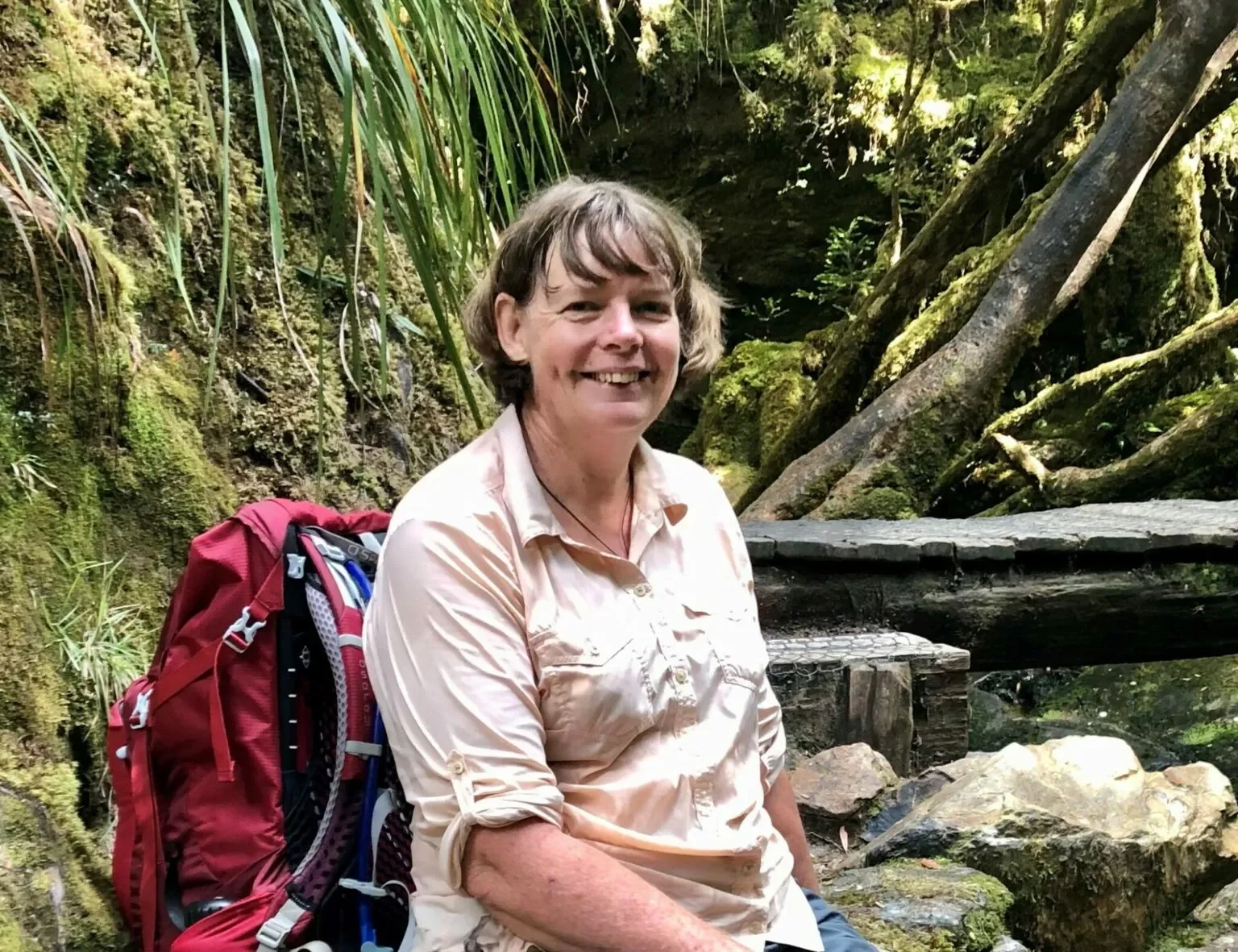A woman with short brown hair smiling, sitting outdoors in a forested area with moss-covered rocks, trees, and ferns around her, wearing a light-colored button-up shirt and a red hiking backpack.