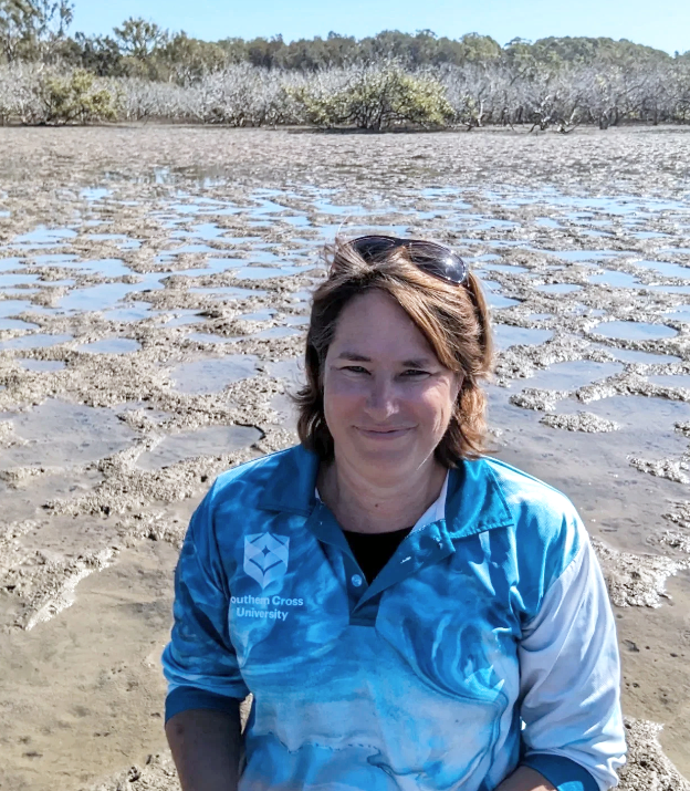 A woman wearing a blue patterned shirt with Southern Cross University logo stands on a sandy, muddy area with puddles, near a body of water with trees in the background.