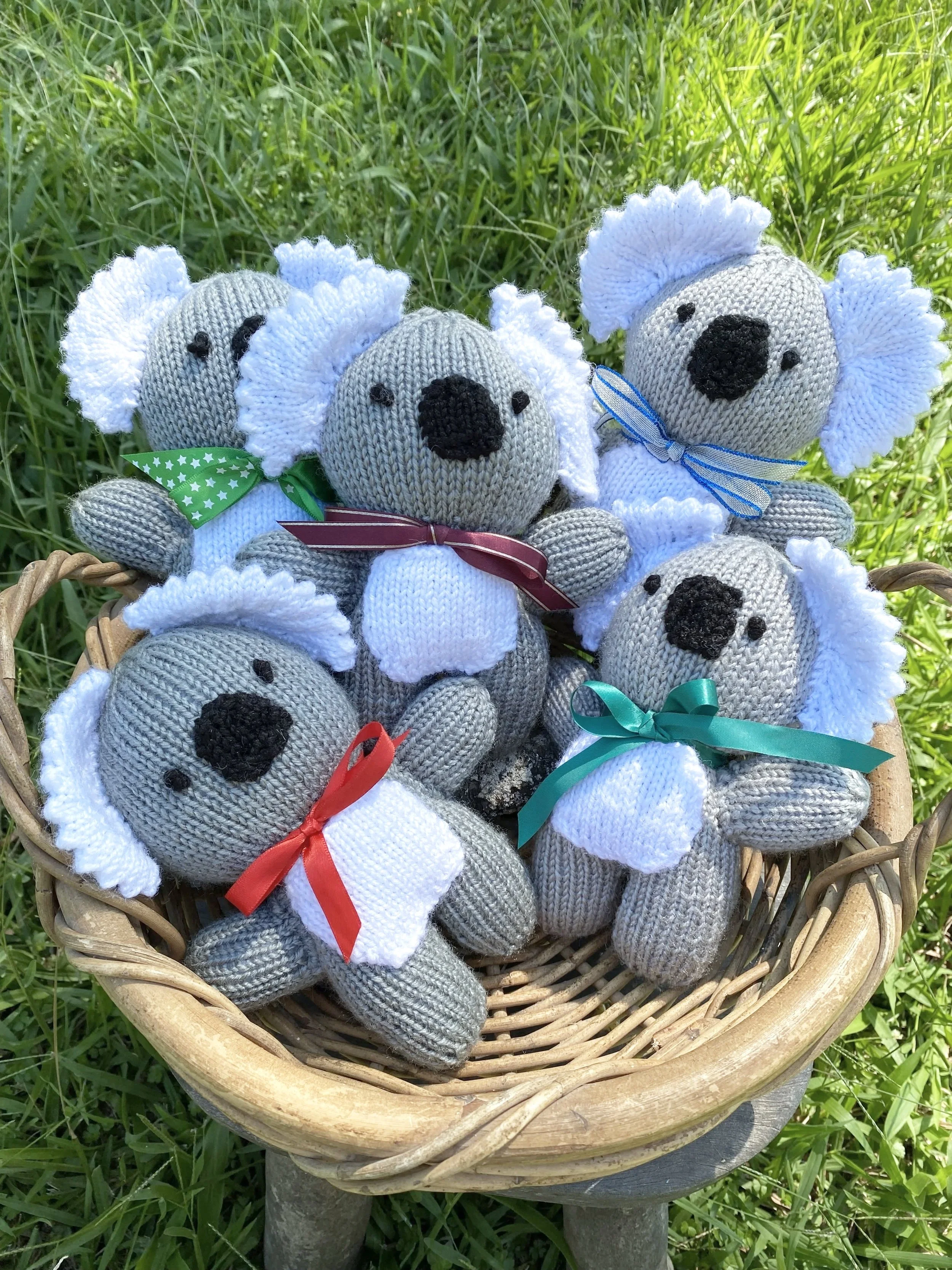 Several handmade knitted koala plush toys with white fluffy ears, black noses, and small black eyes, each with a colorful ribbon around their necks, placed in a wicker basket outdoors on green grass.