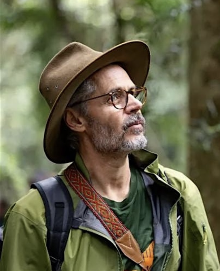 A man is outdoors in a forest, wearing glasses, a wide-brimmed hat, a green jacket, a backpack, and a colorful strap across his chest.
