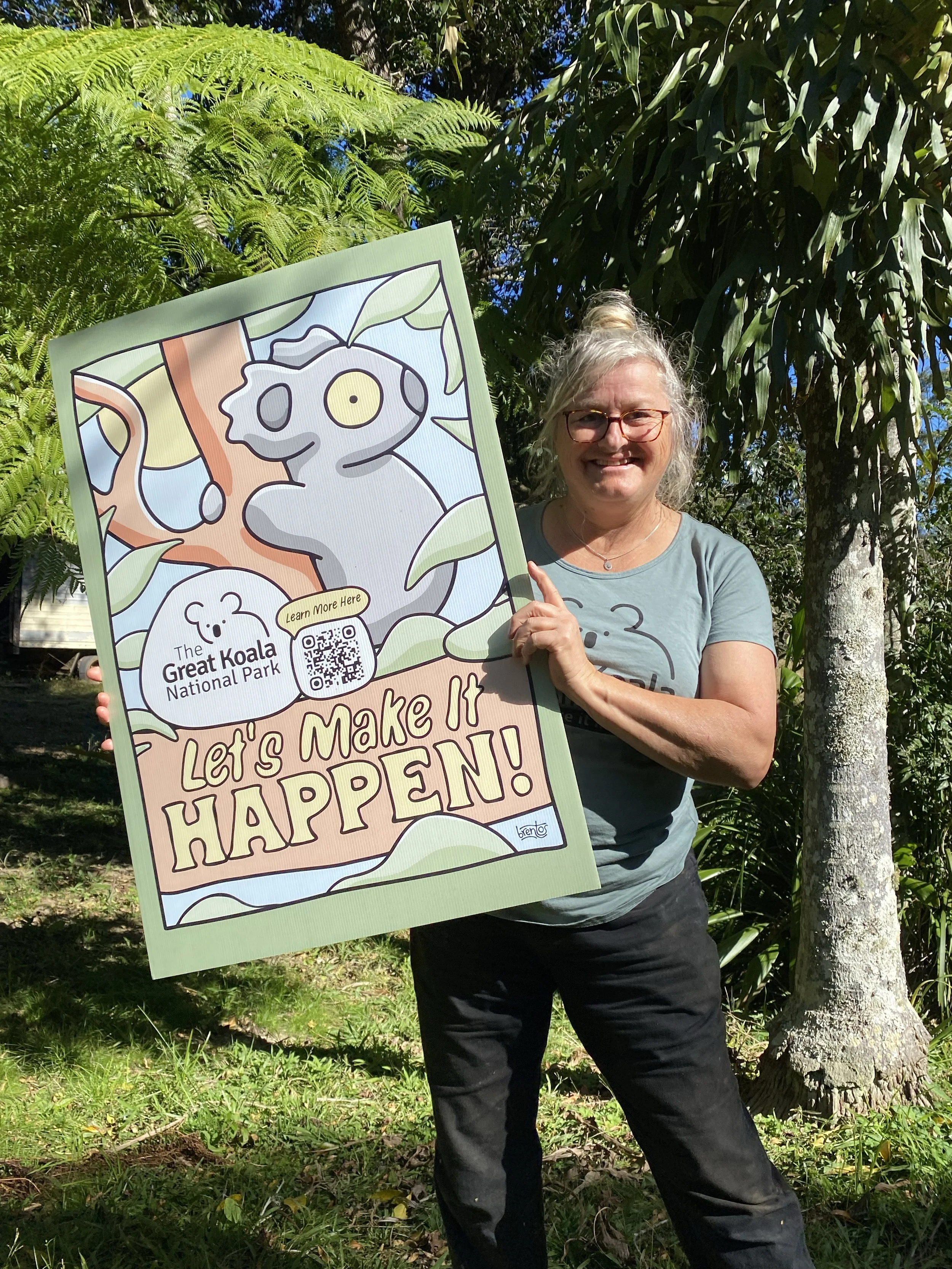 A woman with glasses, gray hair in a bun, smiling, holding a sign promoting the Great Koala National Park, with green trees and grass in the background.