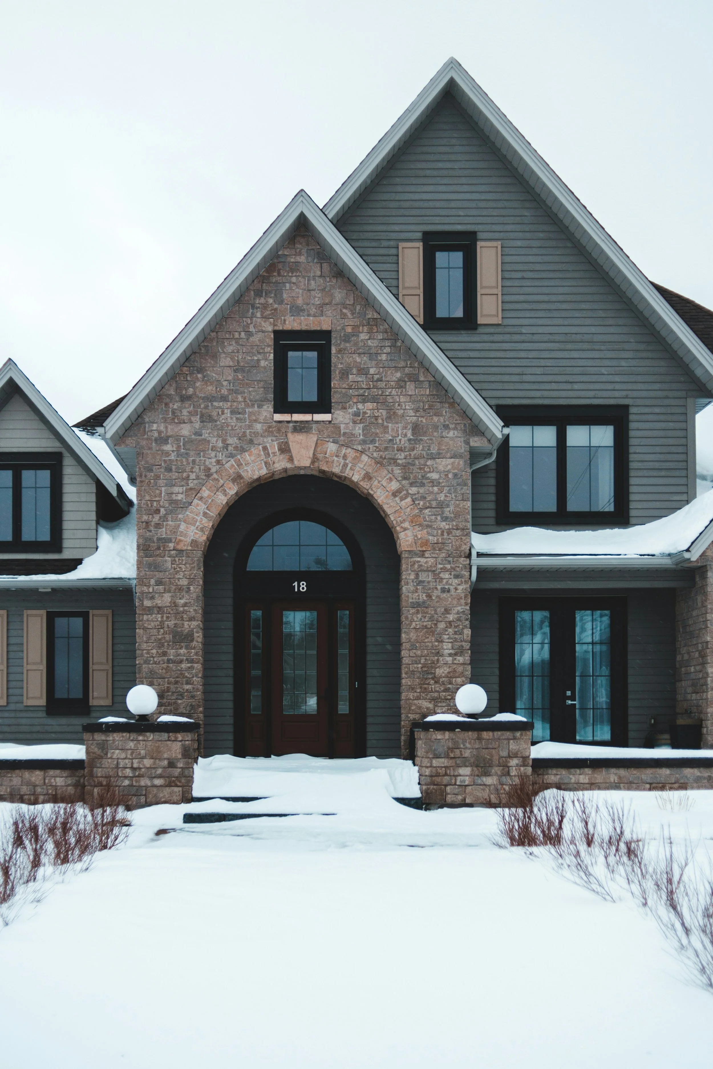 Front view of a modern house with a brick archway entrance, snow on the ground, and snow on the roof, during winter.