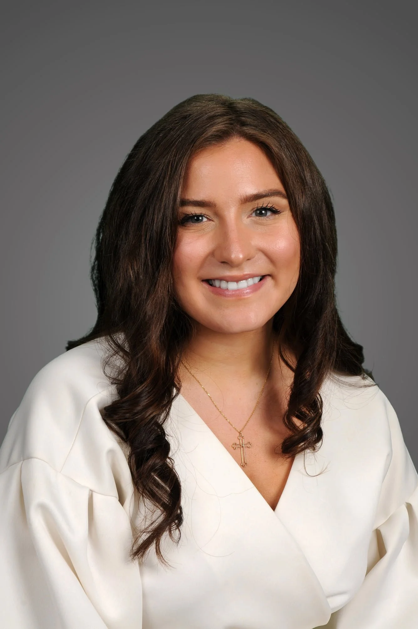 Portrait of a woman with shoulder-length brown wavy hair, wearing a white outfit and a gold cross necklace, smiling against a gray background.