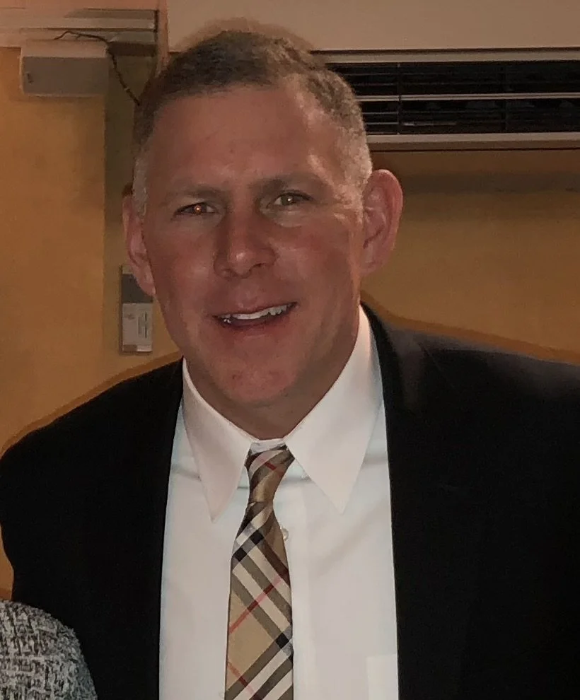 A smiling man wearing a suit, white shirt, and plaid tie, standing indoors with a yellow wall and air conditioning unit behind him.