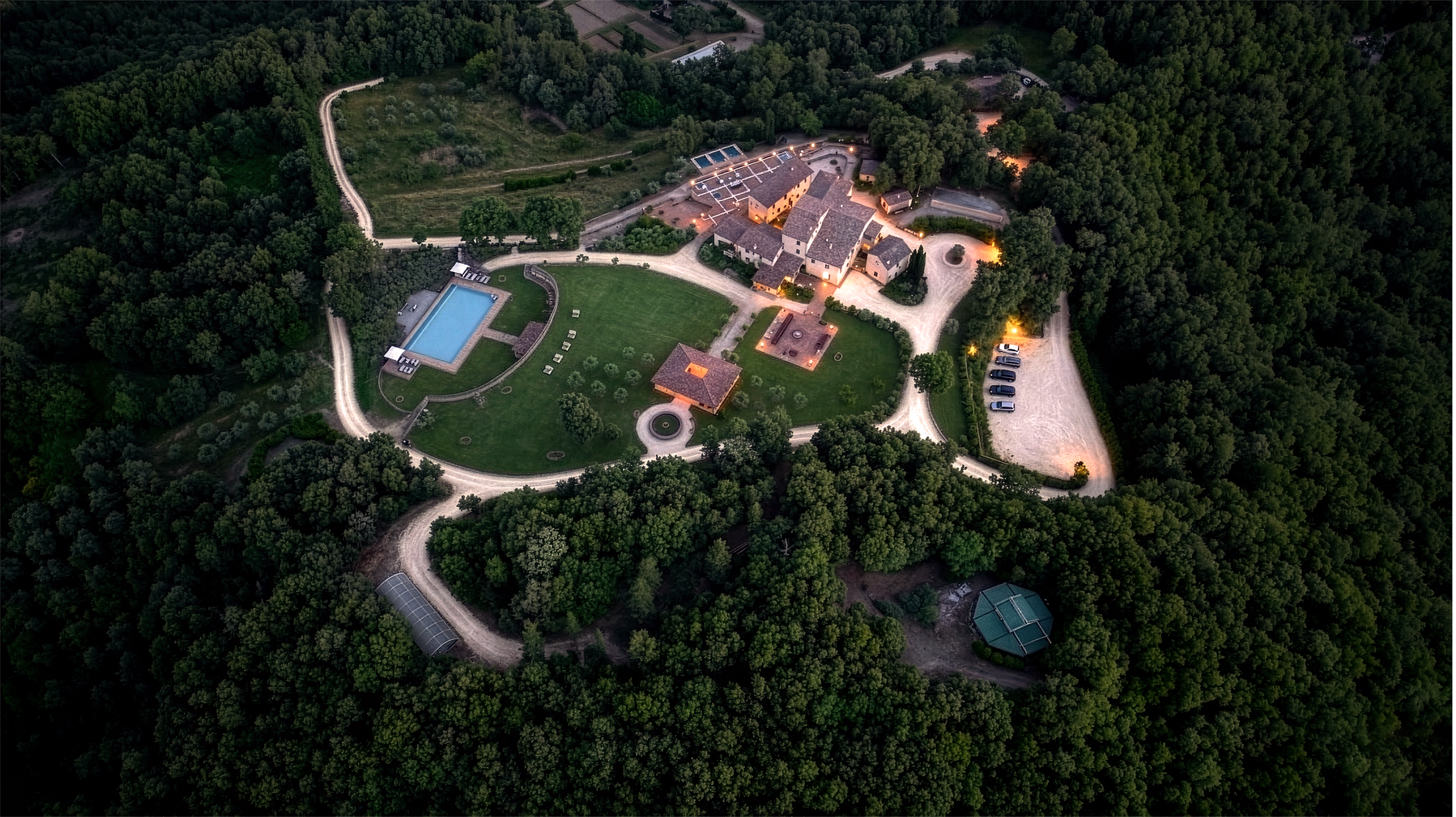 Aerial view of a large estate surrounded by dense forest, featuring a swimming pool, multiple buildings, pathways, and a parking area at dusk.