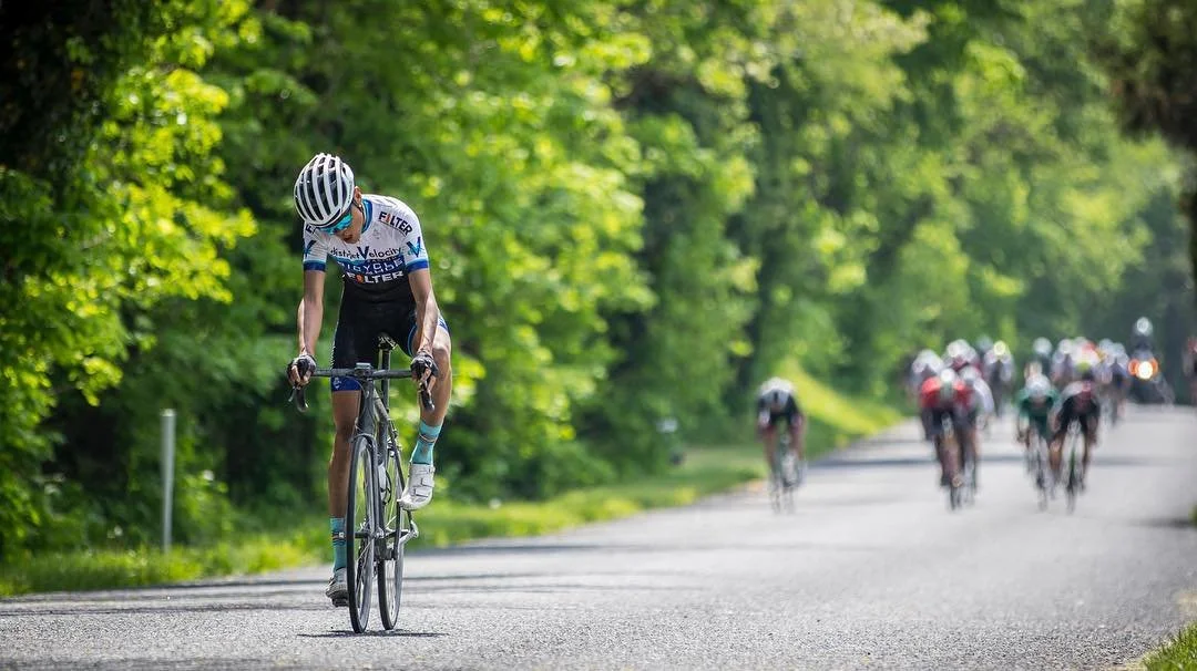 Cyclist wearing a blue and white jersey and helmet riding solo on a road with a group of racers in the background, greenery on both sides, sunny day.