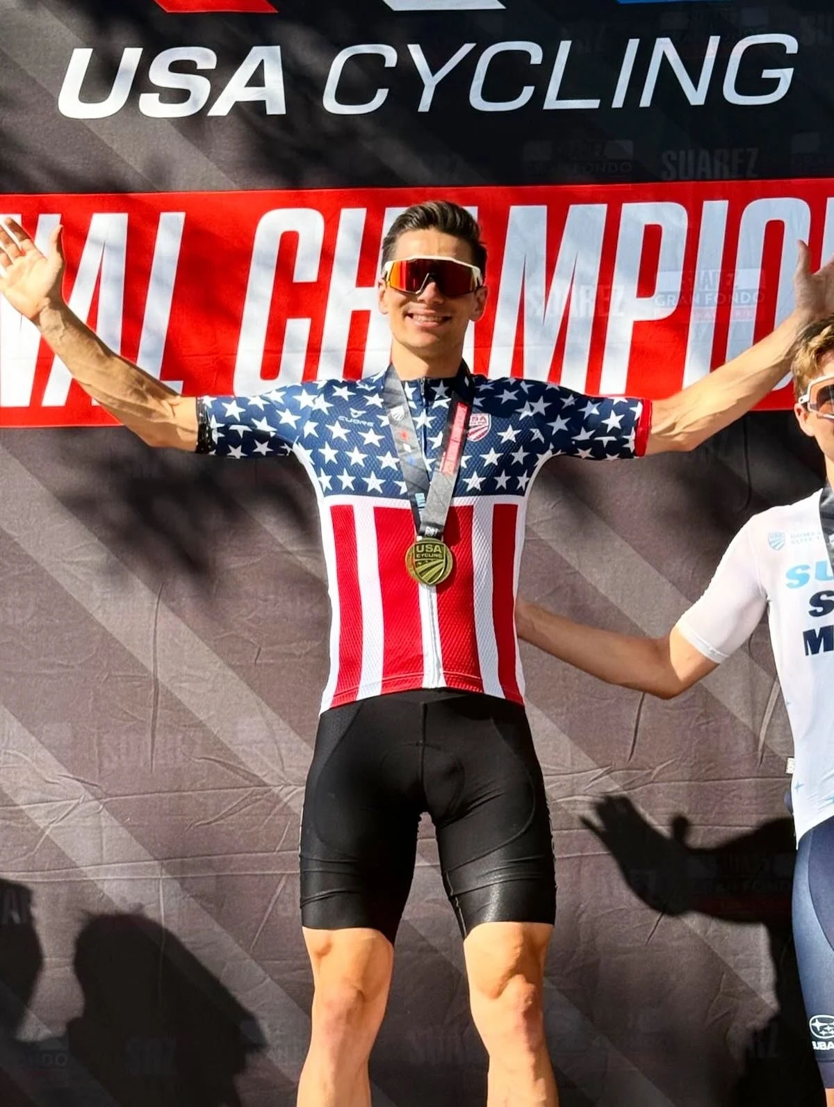 A male cyclist in a patriotic USA cycling outfit with a gold medal around his neck celebrating on a podium.