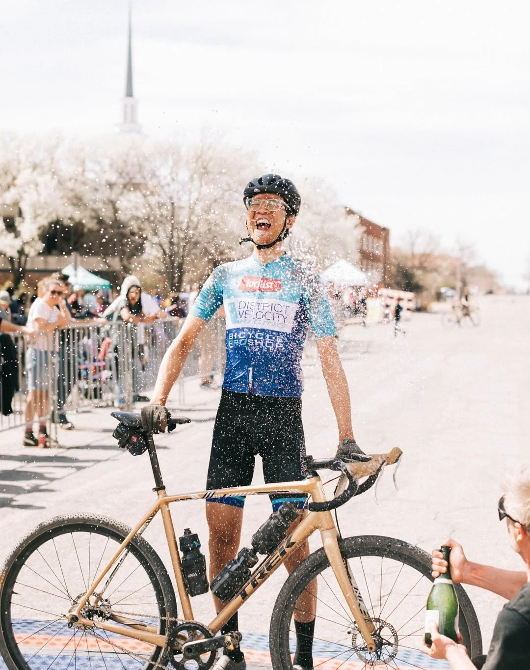 A cyclist happily celebrating at a race event, standing with his bike, while water sprays over him. Spectators are seen in the background.