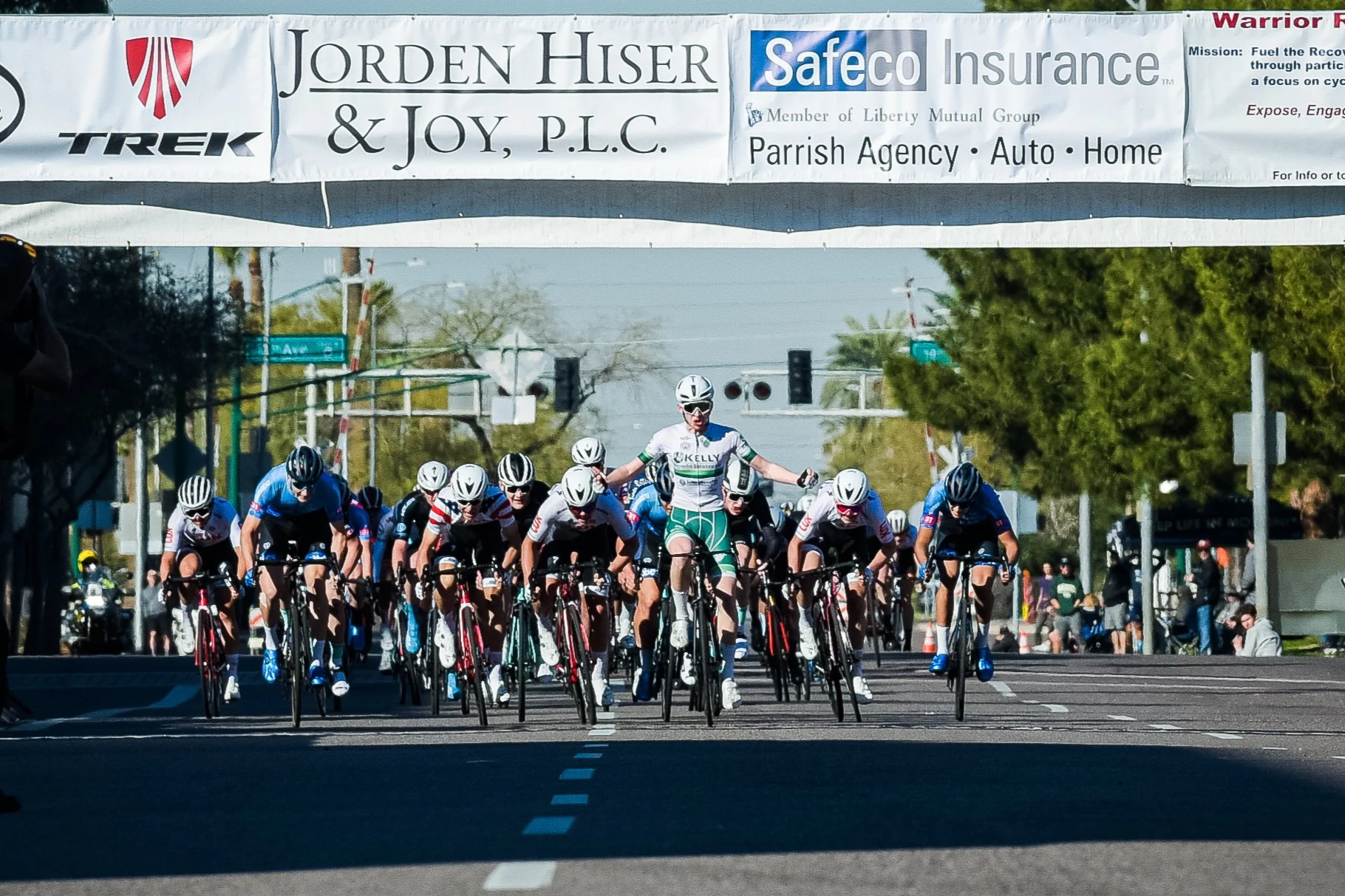 A group of cyclists at the start of a race, riding on a city street under a banner, with some spectators visible in the background.