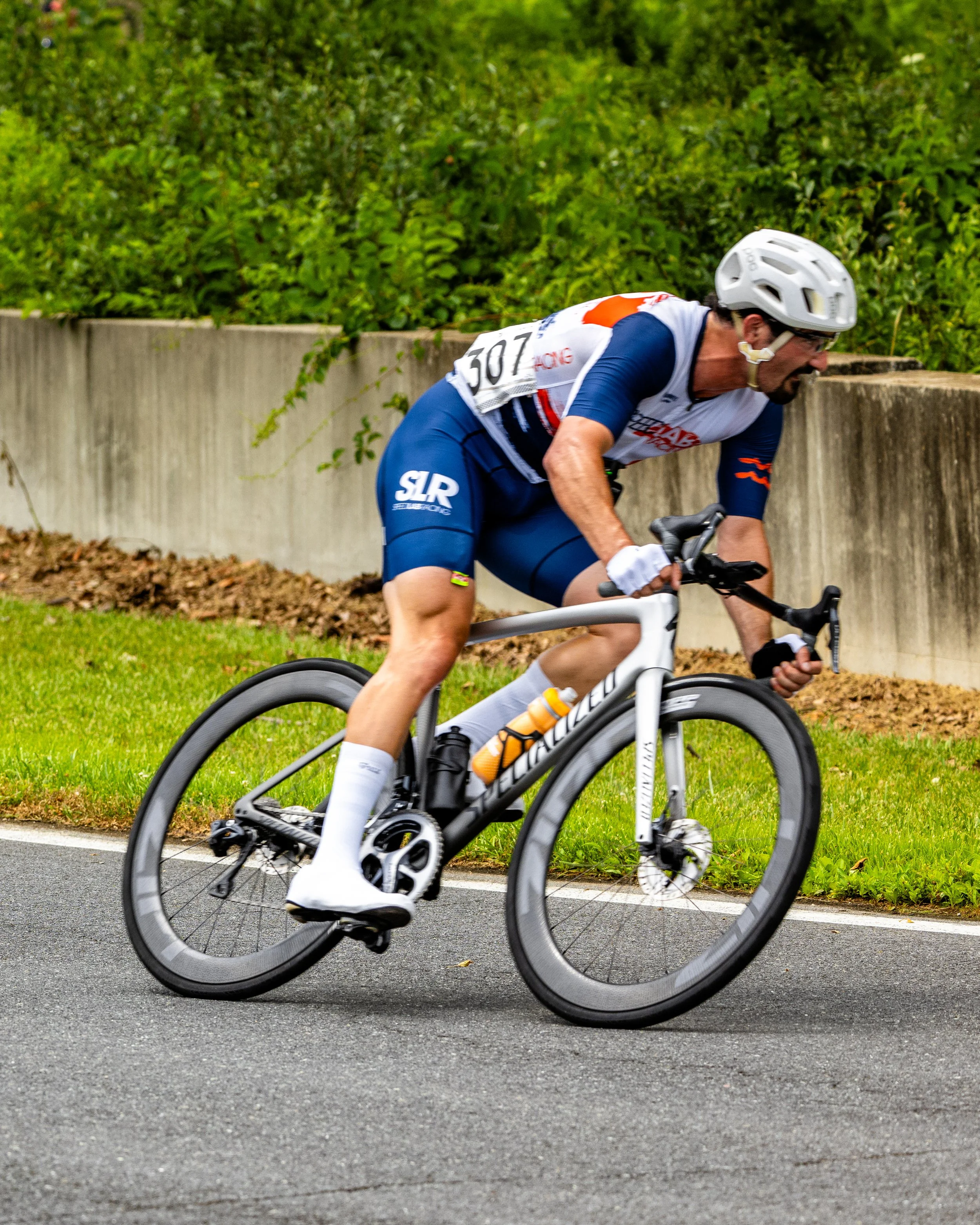 A man riding a gray road bicycle on a paved road, wearing a white helmet, sunglasses, blue cycling shorts, a white and blue cycling jersey, and white socks, with a water bottle attached to the bike.