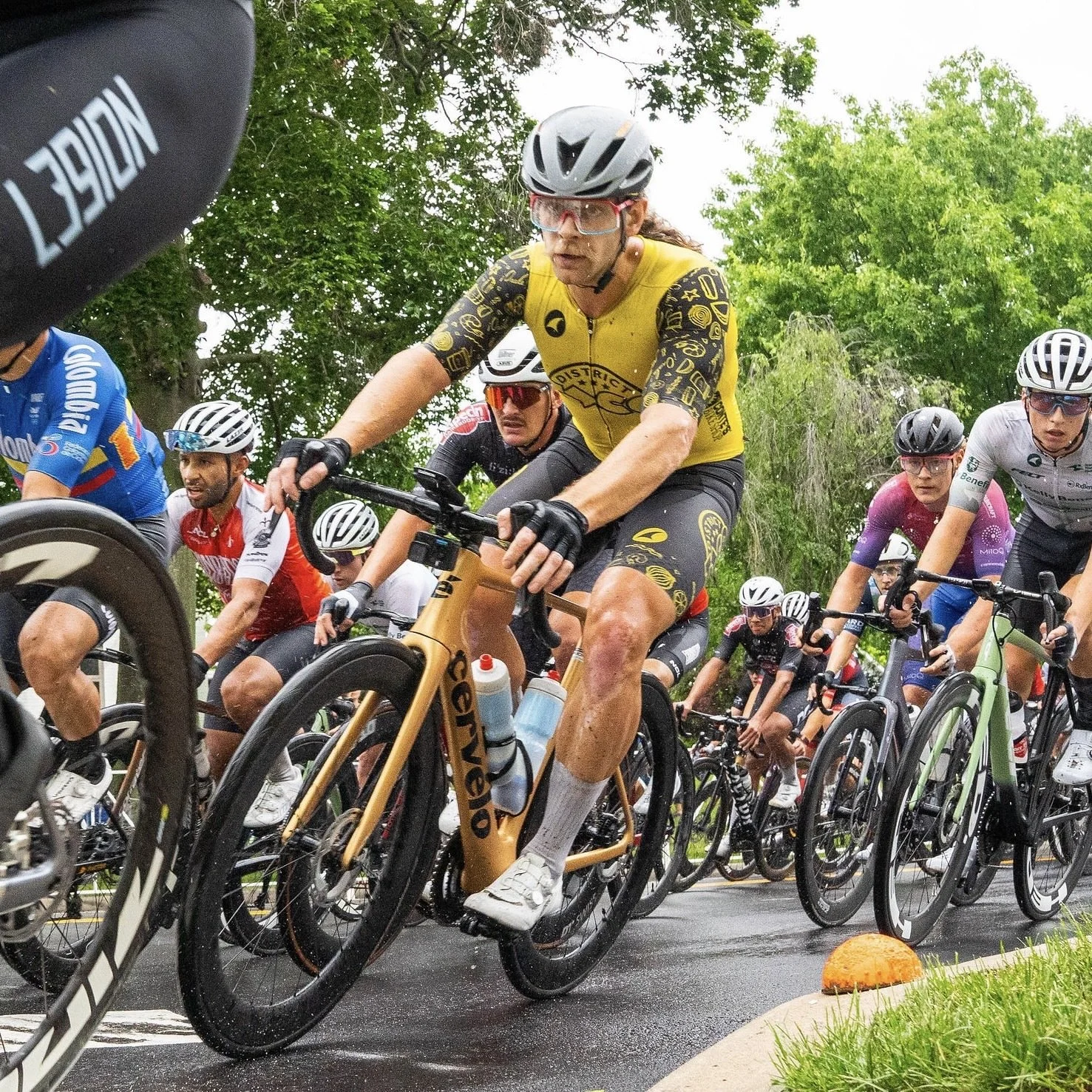 Group of cyclists racing on a wet road surrounded by green trees, with some riders wearing helmets and colorful jerseys.
