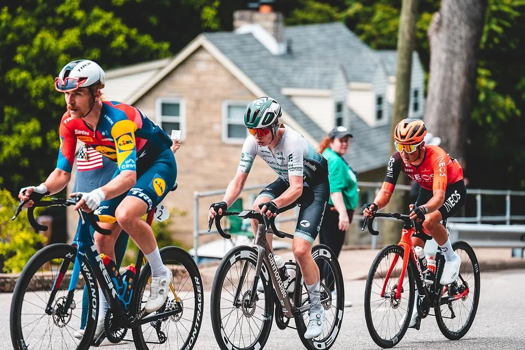 Three cyclists racing on a suburban street with houses and trees in the background.