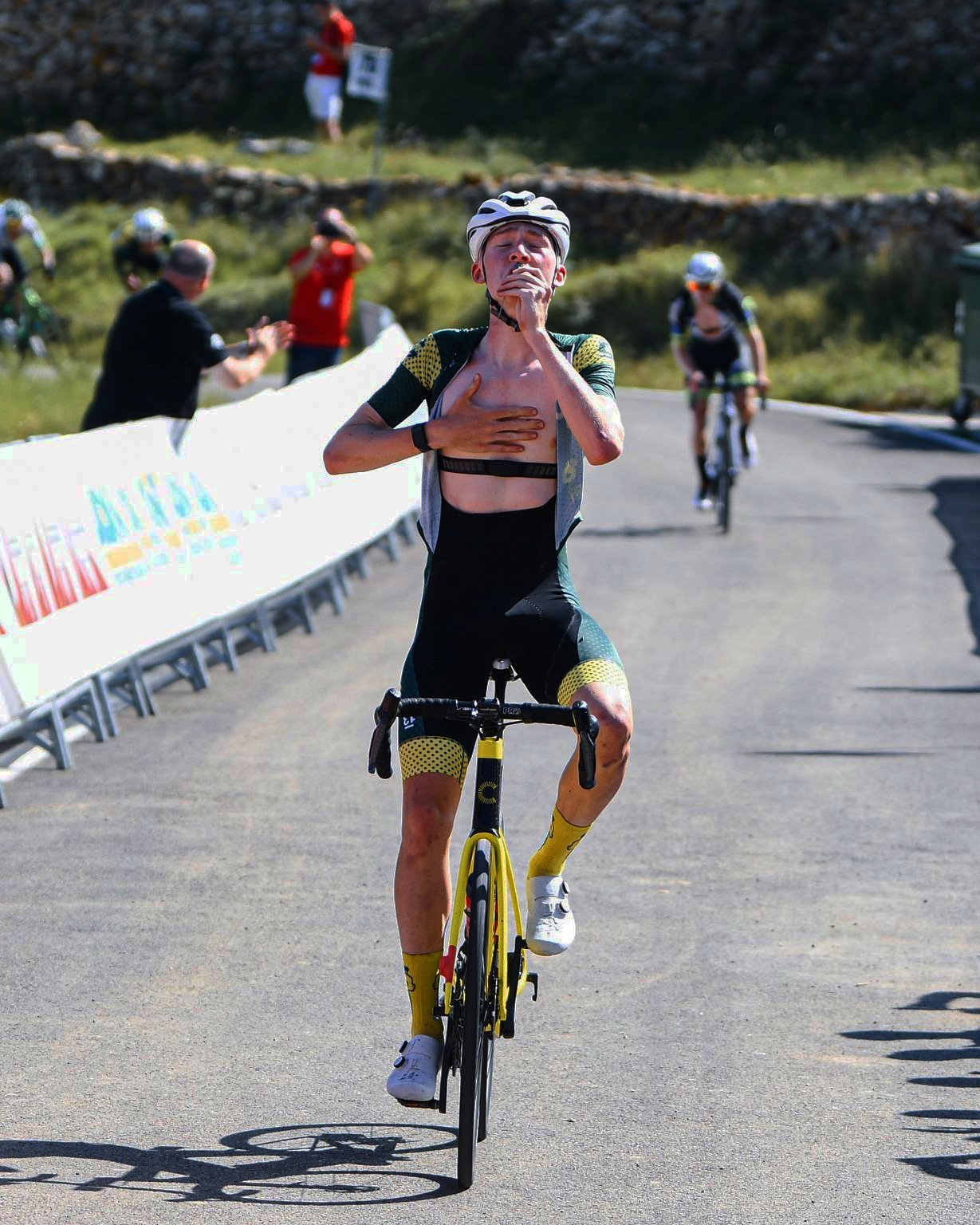 A cyclist in a black and yellow suit with a helmet and yellow socks crossing the finish line on a sunny outdoor road, with other cyclists and spectators in the background.