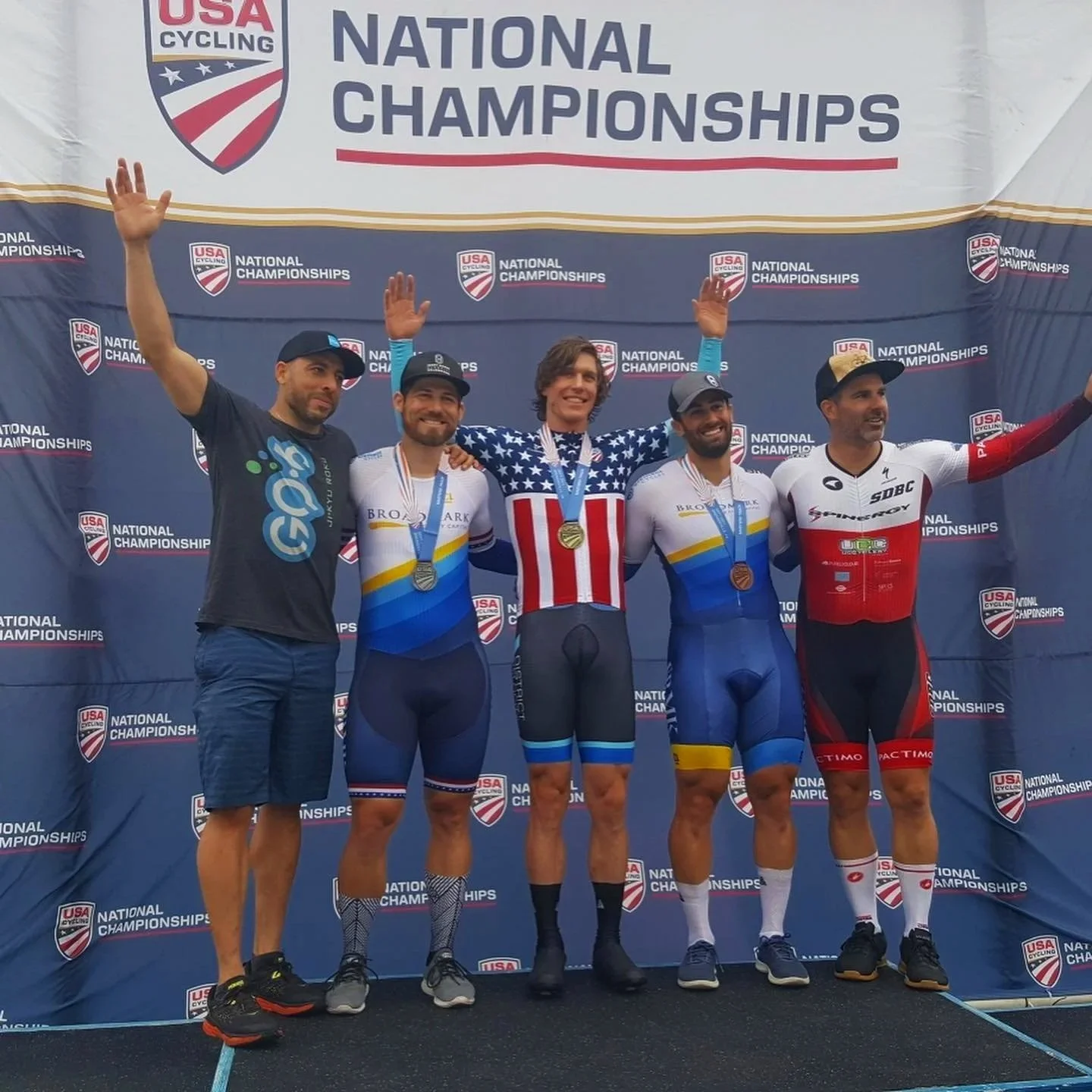 Cyclists standing on a podium celebrating at USA Cycling National Championships. The middle cyclist is wearing a red, white, and blue American flag jersey and holding a gold medal. The other four cyclists wear medals and team jerseys, with two in blue and white, one in red and black, and the fifth in black and blue, smiling and raising their arms in victory.