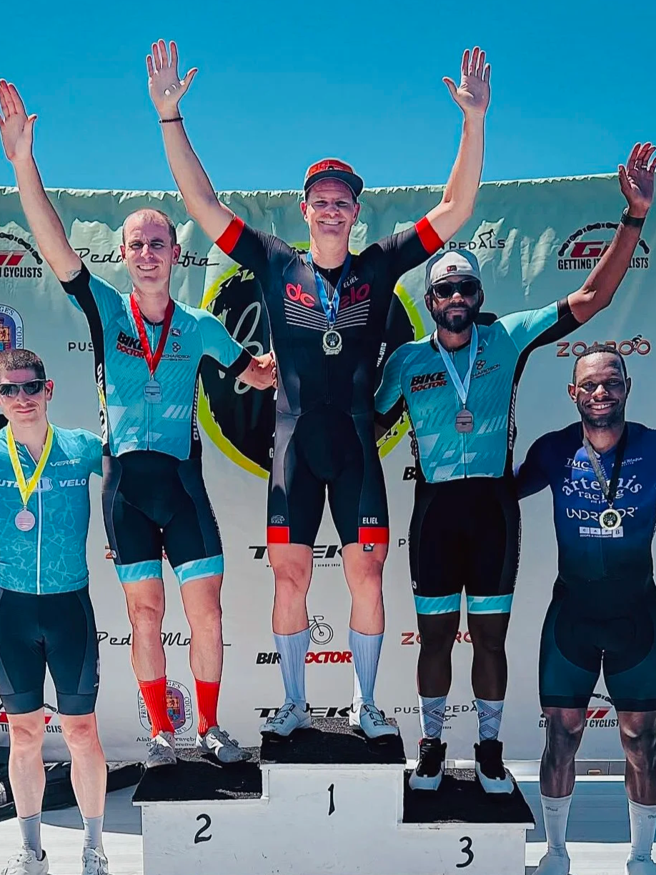 Cyclists standing on a winners' podium celebrating with medals after a race, with the first-place cyclist in the center raising arms in victory.