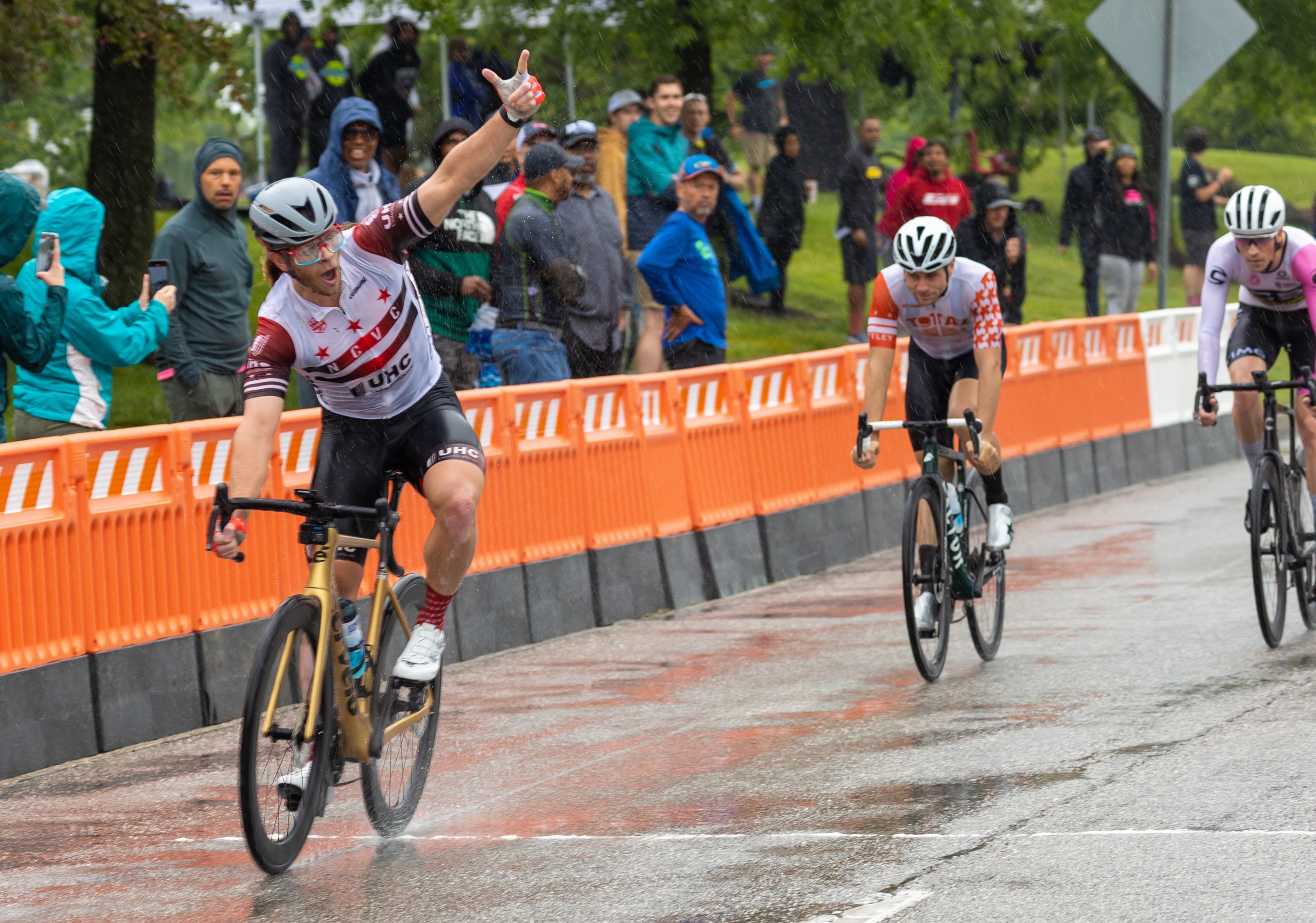 Cyclist celebrating victory as he crosses the finish line in rainy weather during a cycling race, with spectators standing behind orange barricades.