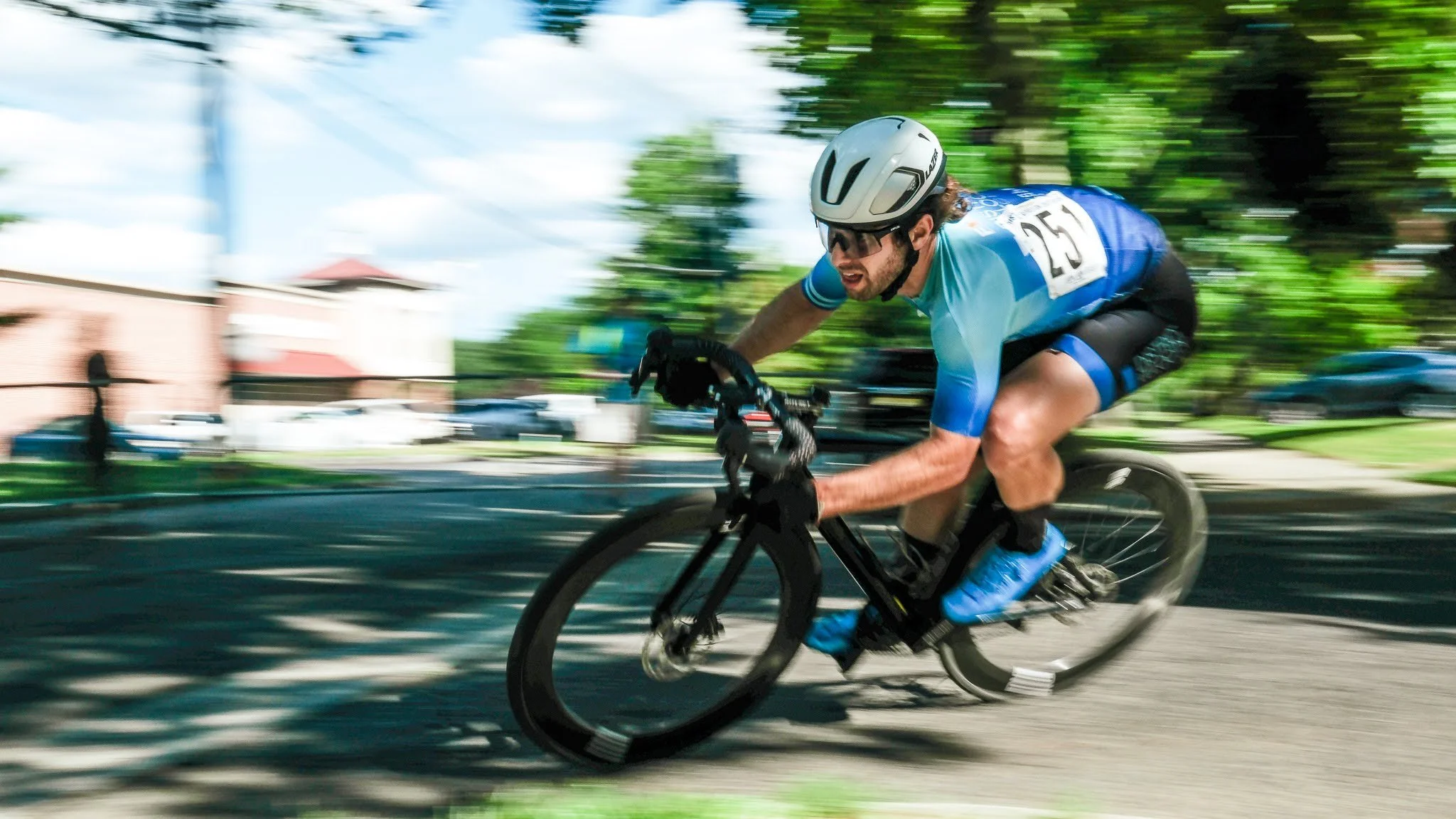 Male cyclist wearing a helmet and sunglasses, riding a mountain bike at high speed on a dirt trail with motion blur from trees and cars in the background.
