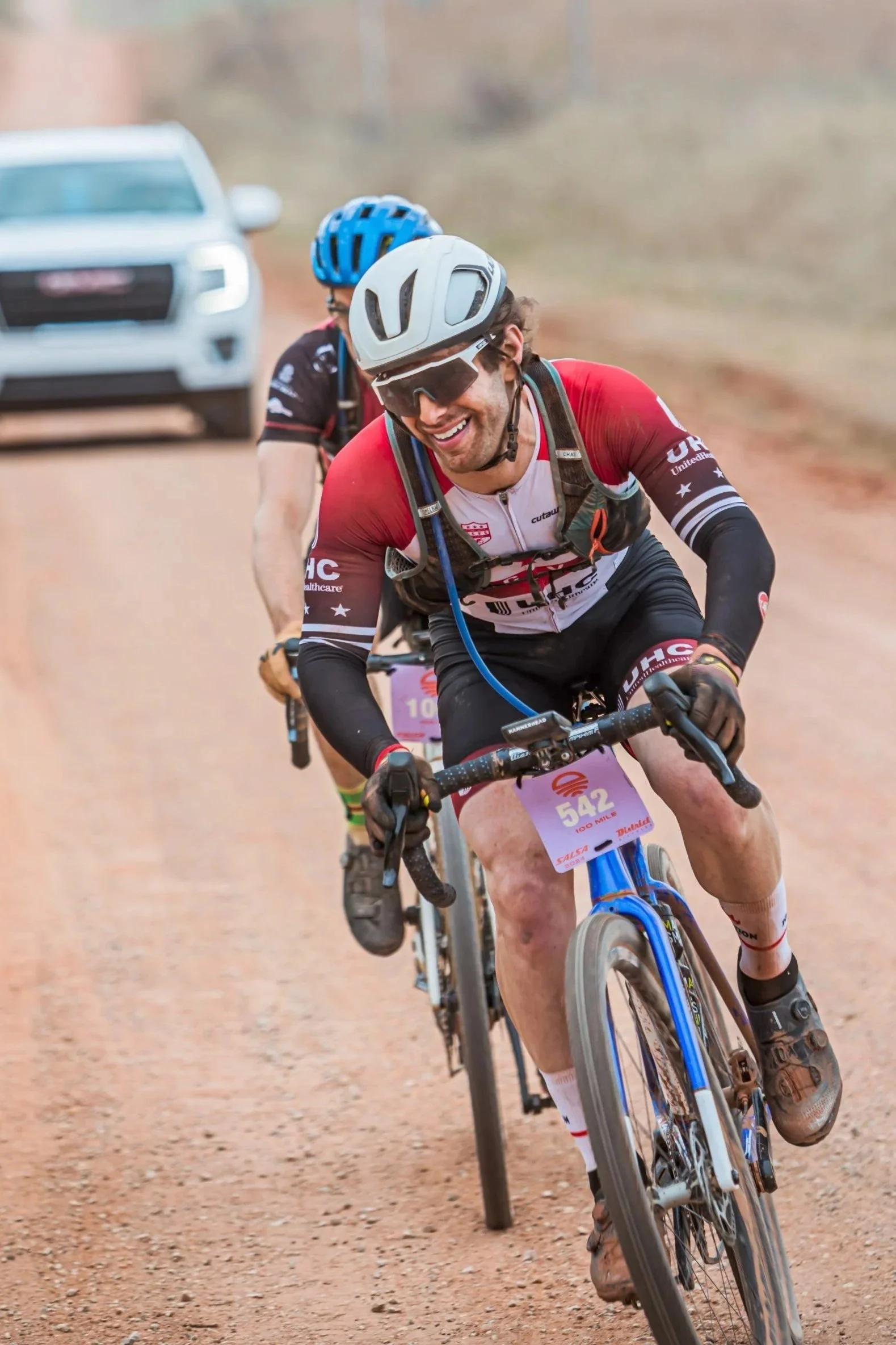Two cyclists riding on a dirt trail during a race, smiling and wearing helmets and race gear, with a vehicle in the background.