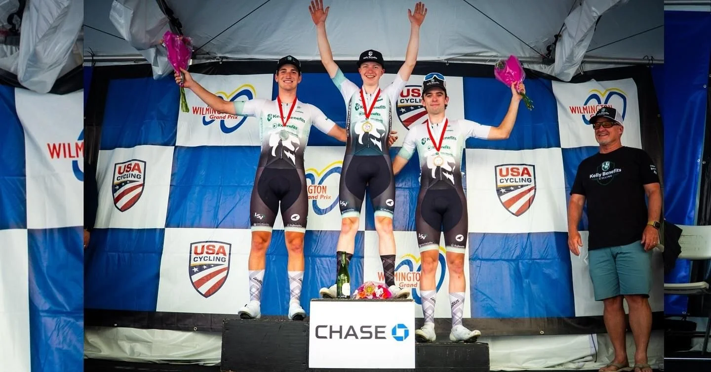 Three male cyclists in race uniforms standing on a winners' podium with medals, holding pink bouquets, celebrating with a man in shorts and a T-shirt, under a tent with banners that say 'USA Cycling' and 'Wilmington Grand Prix'.