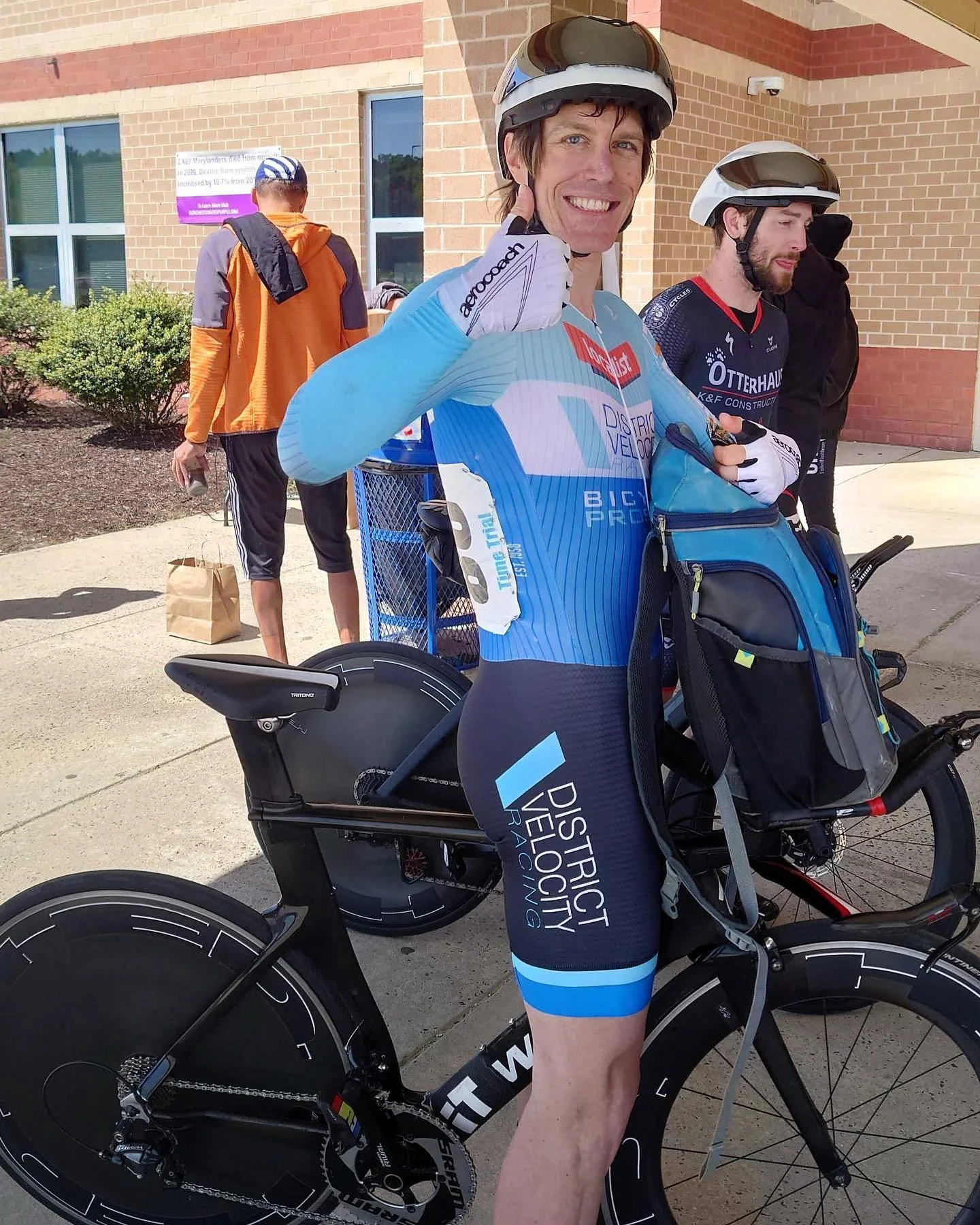 A cyclist in a blue and black racing suit giving a thumbs-up beside his black specialized bicycle, with two other cyclists in helmets and gear in the background outside a brick building.