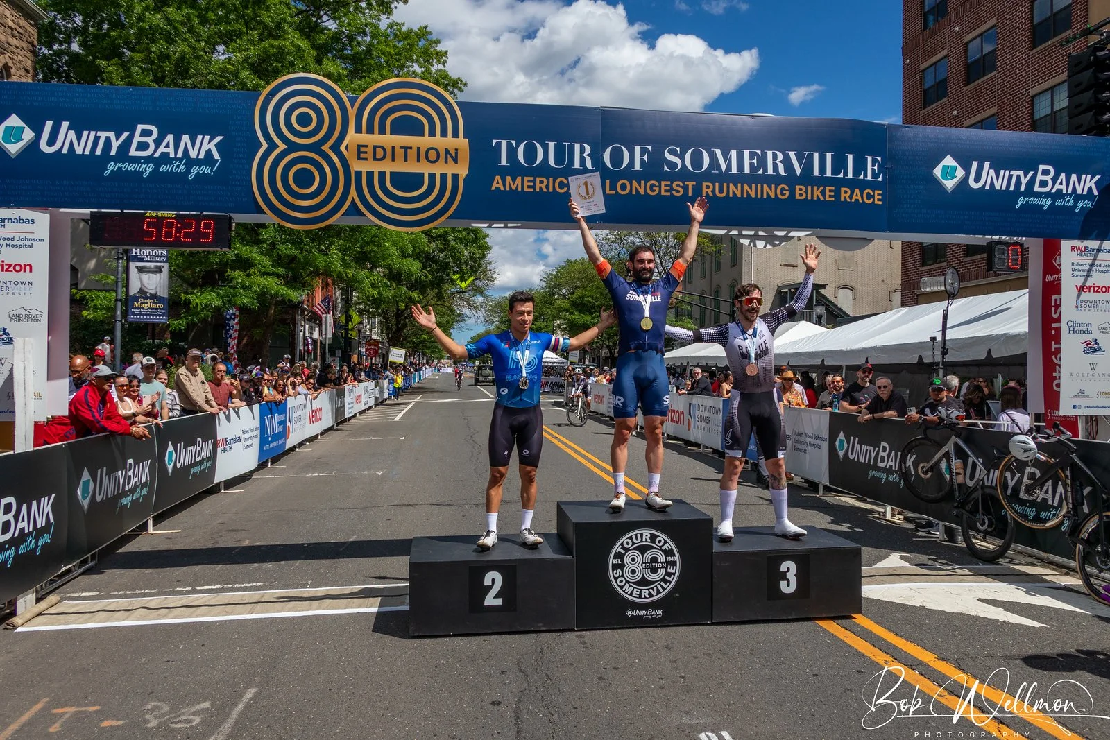 Three male athletes on a winners' podium at a cycling race event. The first-place winner is in the center, wearing a blue outfit, with his arms raised and a medal around his neck. The second-place finisher is on the left, also in a blue outfit, with his arms out. The third-place finisher is on the right, dressed in black and white, also with his arms raised. The background features a street lined with spectators, banners, and tents, with a large arch overhead displaying the event's name and sponsors.