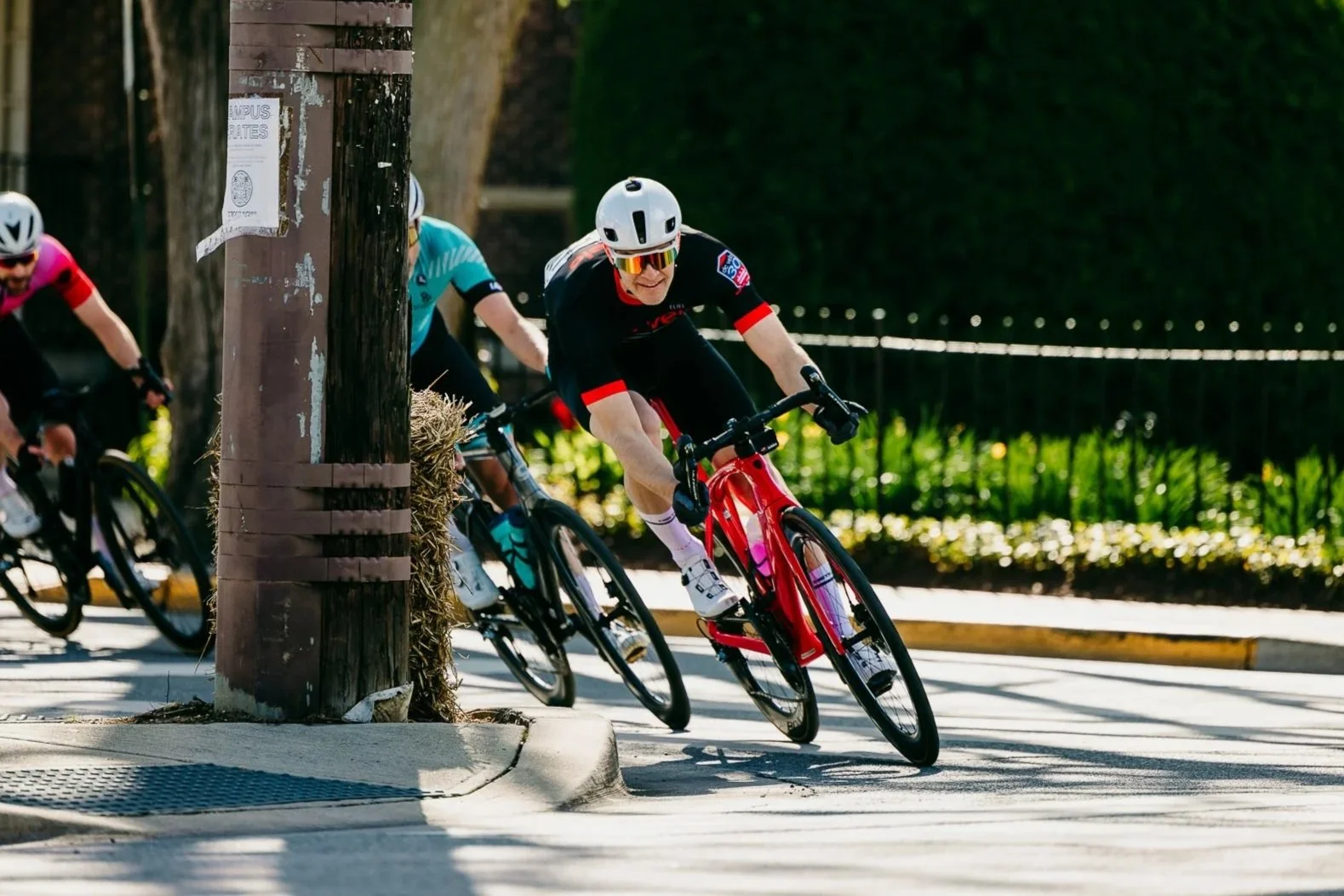 Cyclists racing on a sunny road, leaning sharply around a corner with a utility pole in the foreground and green plants in the background.