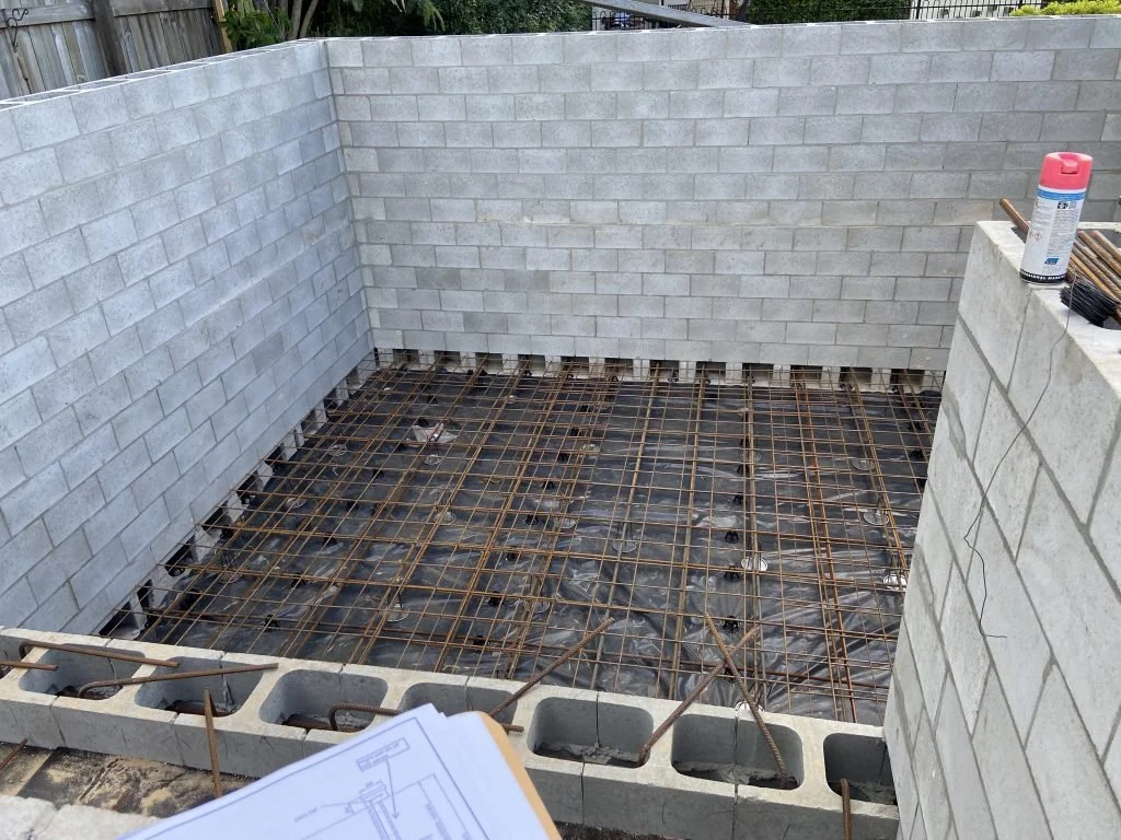 Construction site with cinder block walls and a rebar grid for a concrete foundation or floor, with some construction tools and materials visible.