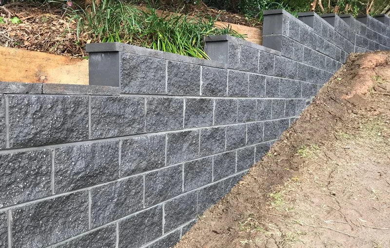 A gray stone retaining wall with cap stones, built on a slope with soil and plants above it.