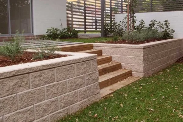 A landscaped backyard with a raised garden bed made of light-colored concrete blocks, wooden steps leading up to it, and some greenery and plants inside.