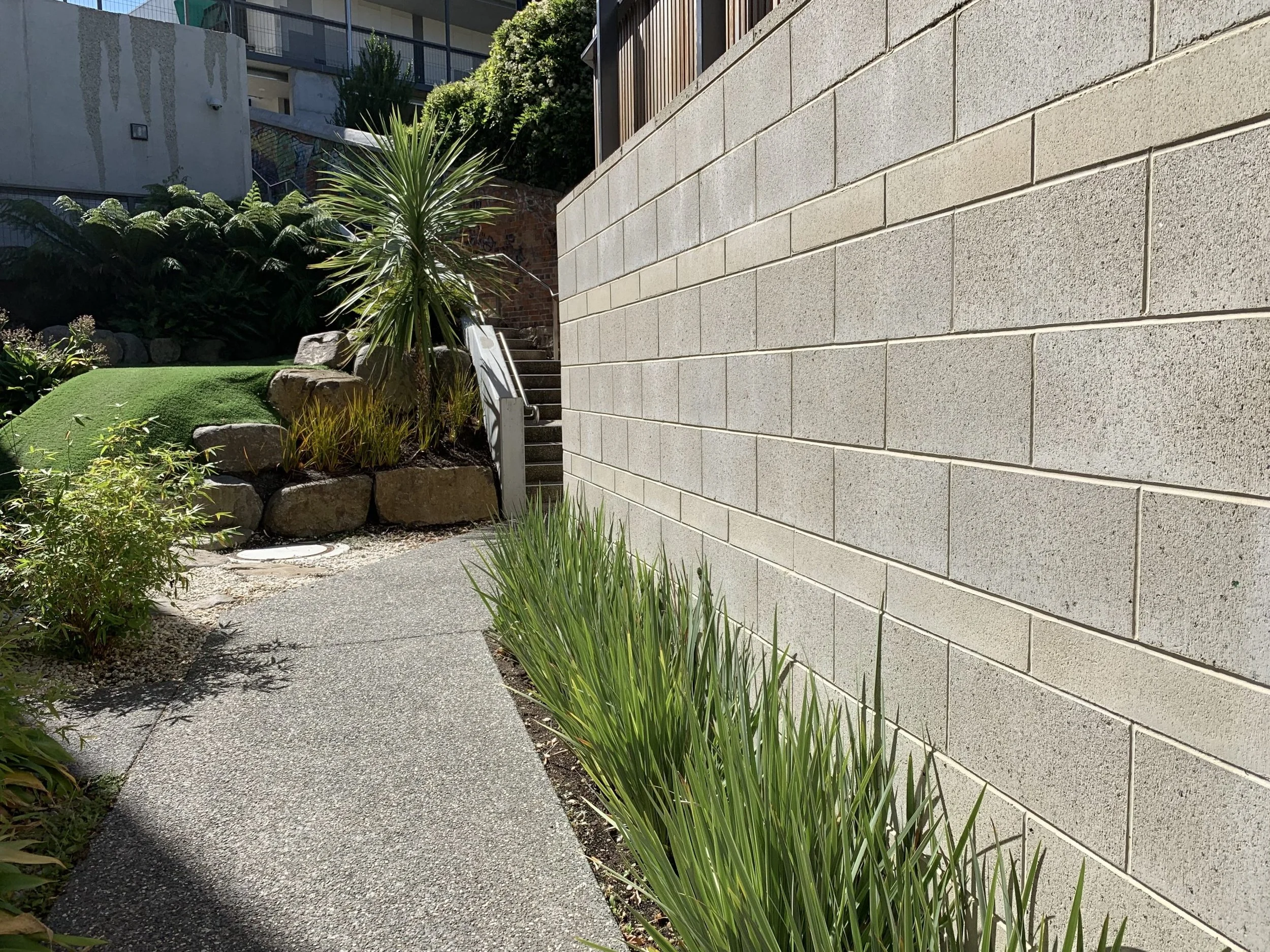Pathway beside a beige brick wall with green plants and bushes on the left side and stairs with a metal railing leading up to a garden area.