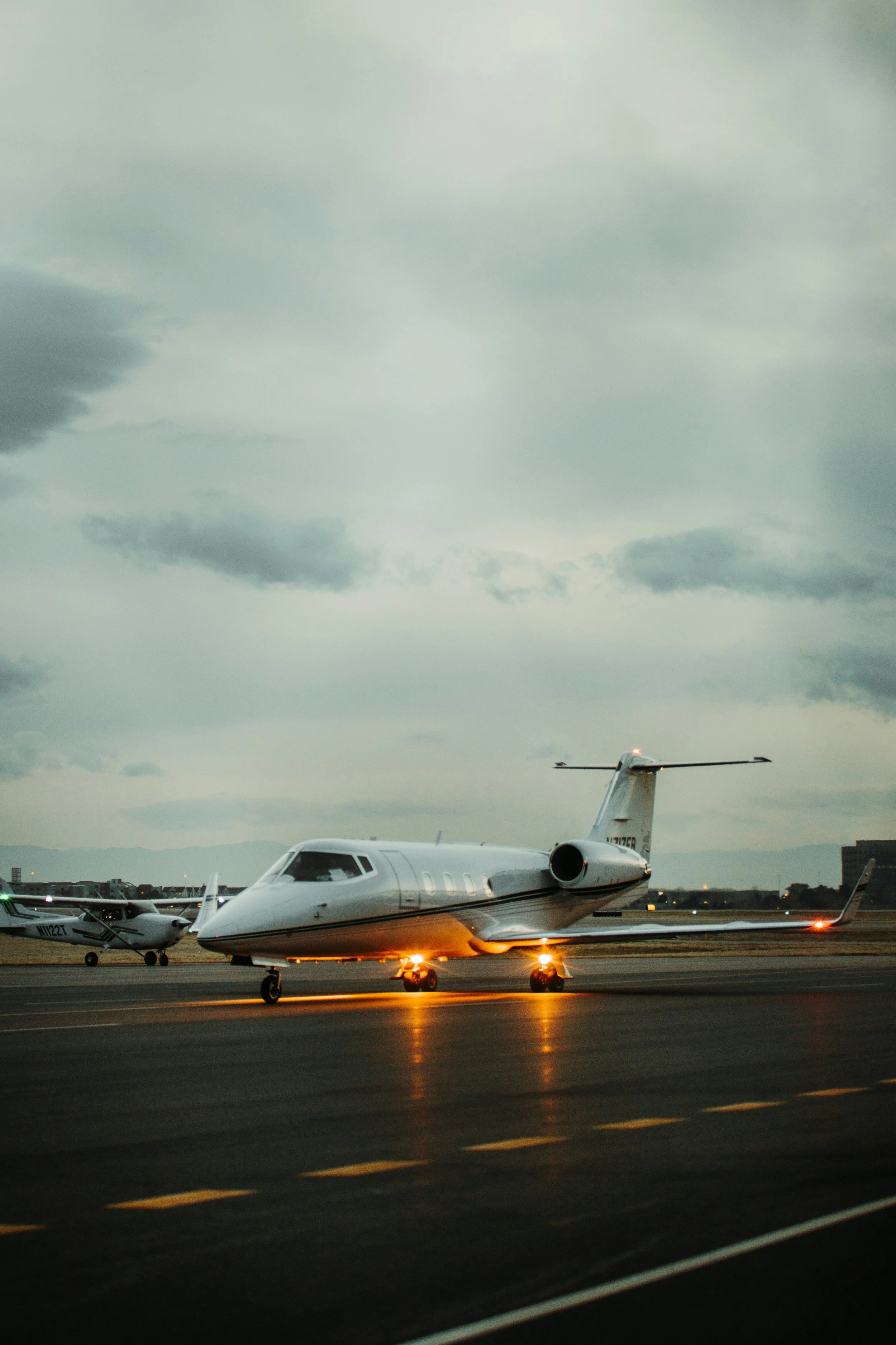 Private jet taxiing on an airport runway during dusk with cloudy skies, surrounded by other small aircraft and cityscape in the background.