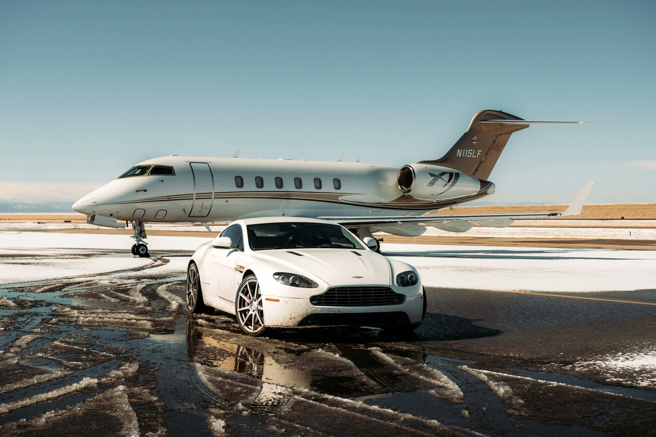 A white luxury car parked on wet, snowy tarmac in front of a private jet at an airport.