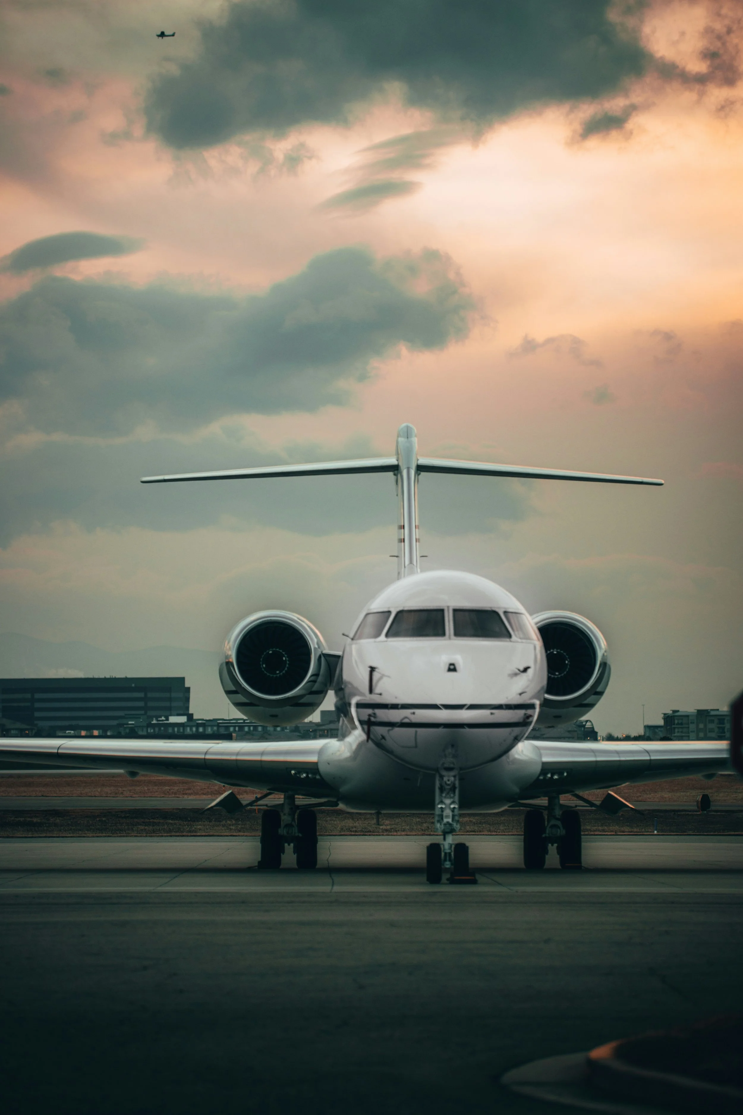 Front view of a private jet on an airport tarmac with a cloudy sky and an airplane flying in the background at sunset.