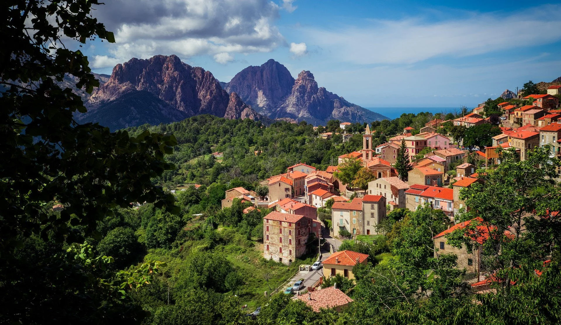 Scenic view of a hillside village with terracotta-roofed houses, surrounded by green trees and mountains, under a partly cloudy sky.