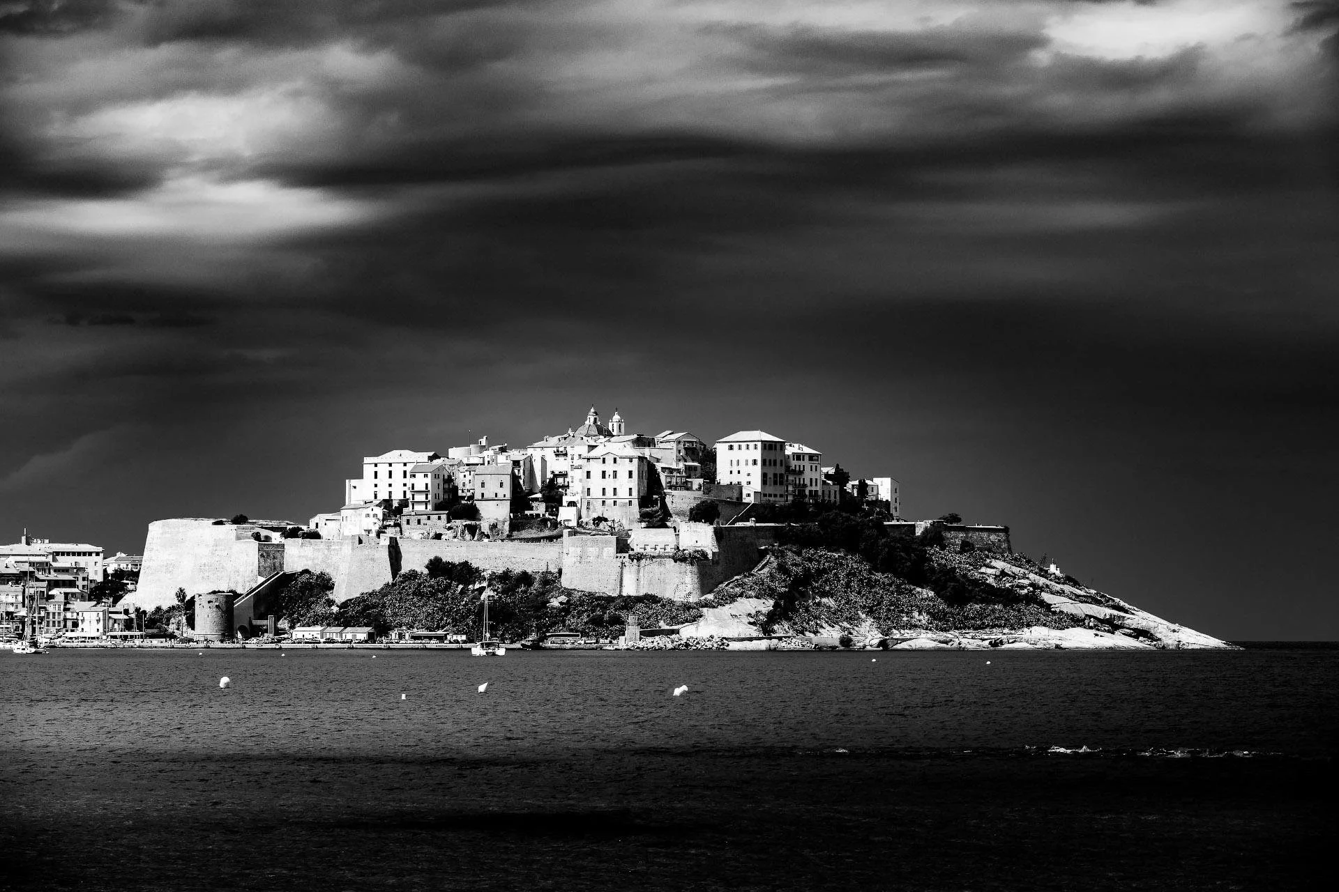 Black and white photo of a castle or fortress on a hill overlooking water, with dark stormy clouds overhead.