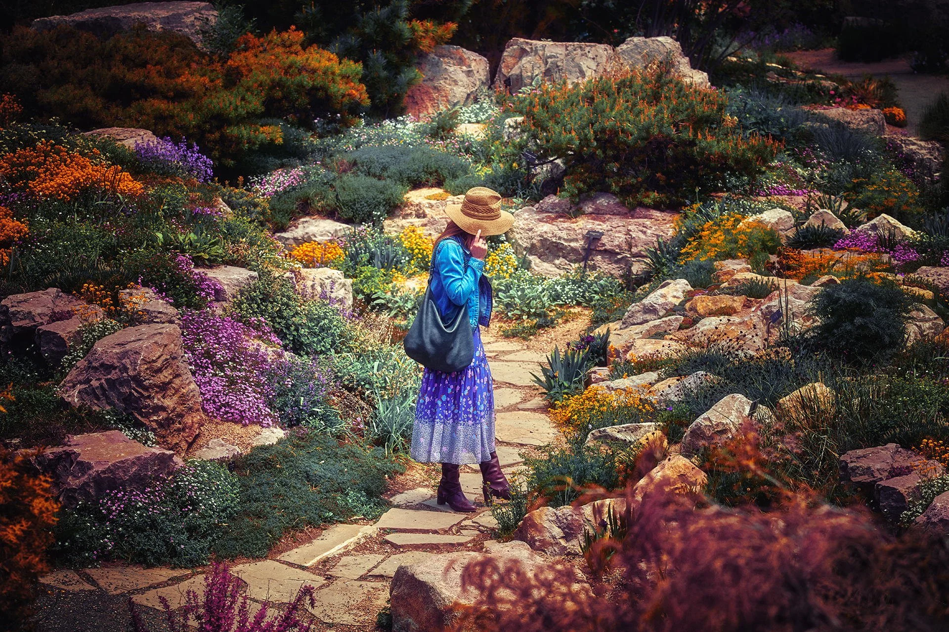 A woman in a blue jacket and a purple dress with white patterns, wearing a wide-brimmed straw hat, walking on a stone pathway through a colorful garden with rocks, various green plants, and blooming flowers of pink, purple, yellow, and orange during 