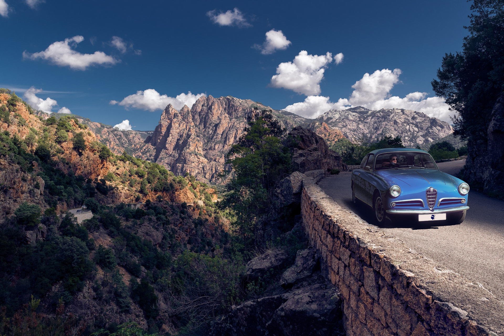 A vintage blue car parked on a winding mountain road with rocky hills and green trees under a partly cloudy sky.
