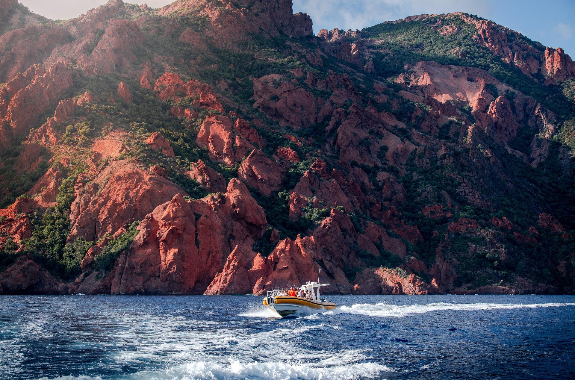 A boat sailing on water with red and brown rocky cliffs and green vegetation in the background.