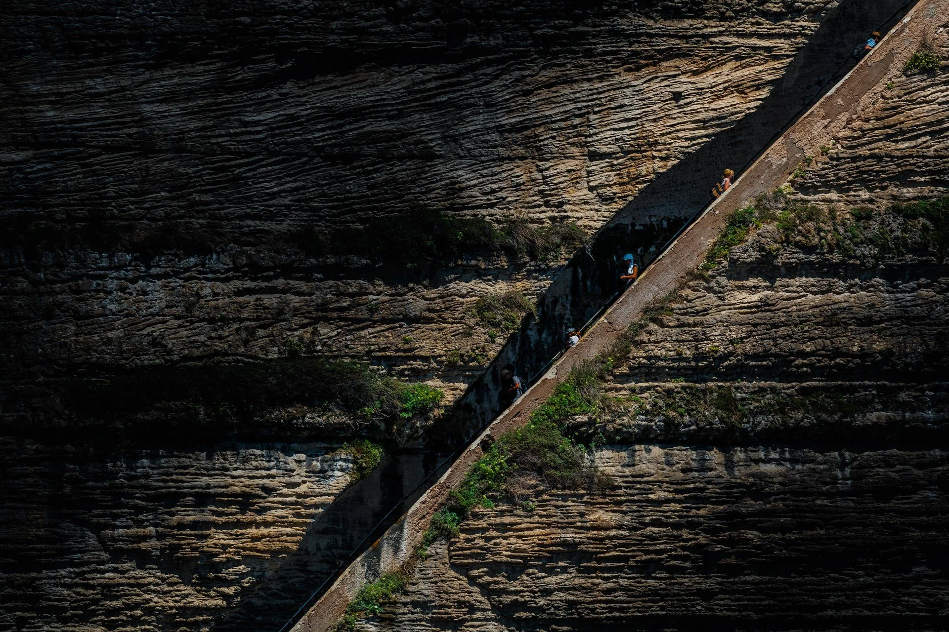 People walking on a steep, narrow concrete path built into a rocky cliffside.