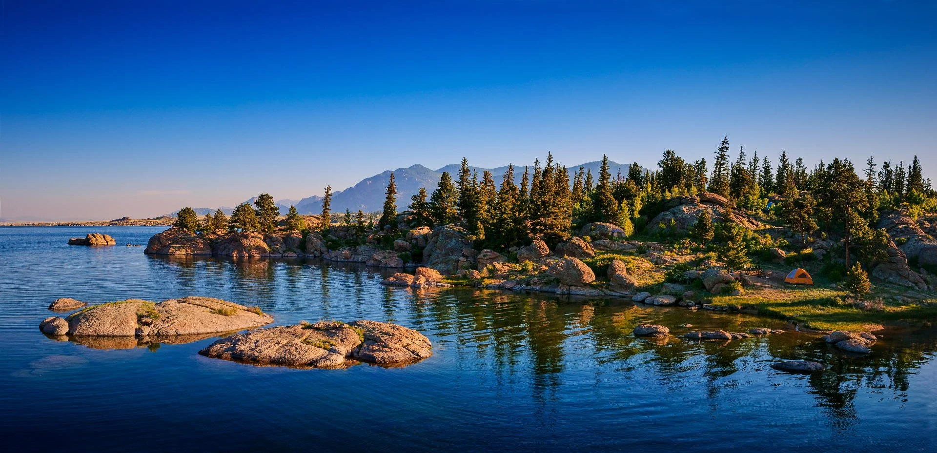 A scenic lakeside view with calm blue water, large rocks, a forest of pine trees, and mountains in the background under a clear blue sky.