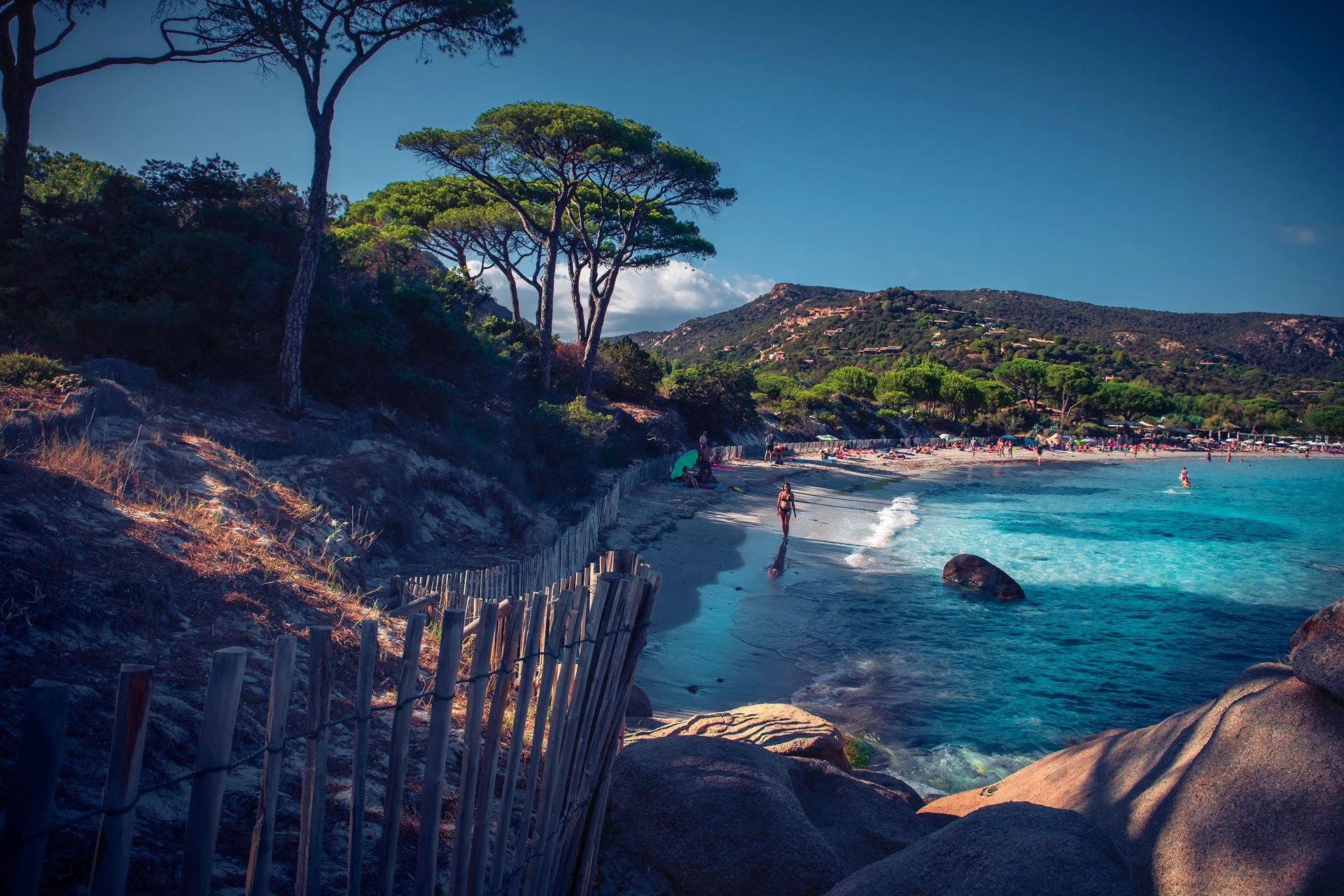 A scenic beach with clear blue water, sandy shoreline, rocks, and trees on a sunny day with a few people walking and relaxing.