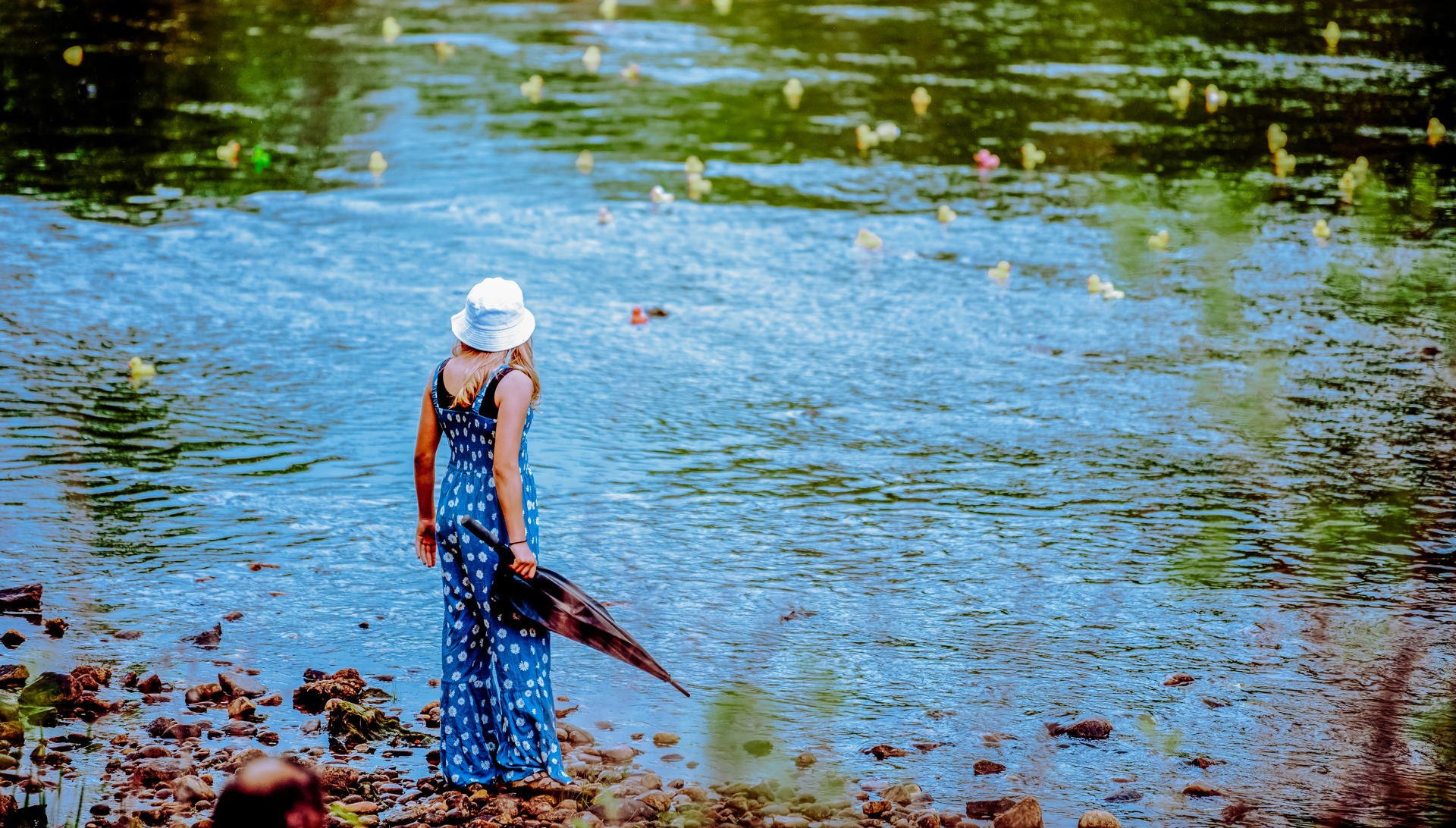 A girl in a blue dress and white hat stands at the edge of a river, holding a black umbrella, watching a rubber duck race.