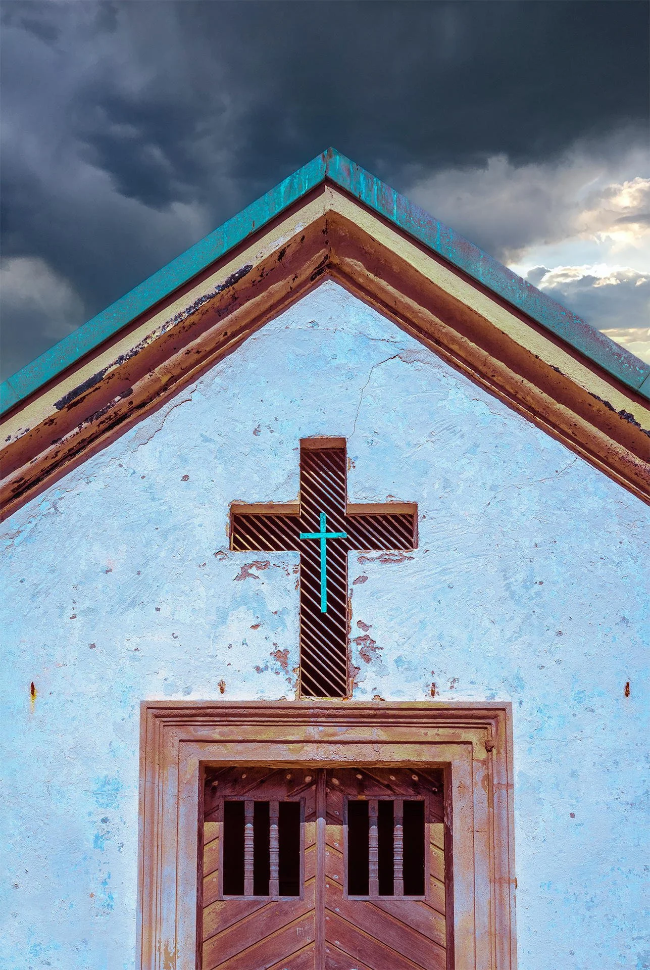 Front of a church with a wooden door and a cross-shaped window above it, weathered white exterior wall, dark stormy sky in background.