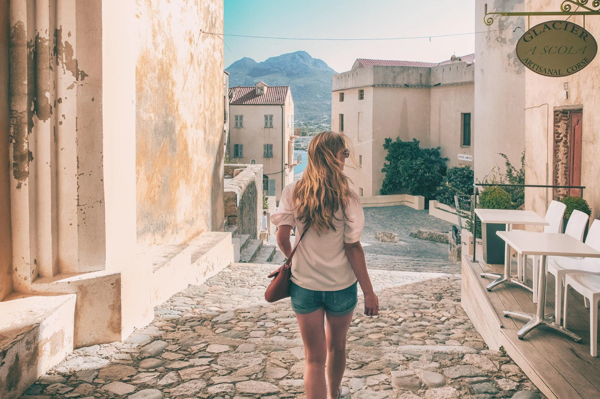 A woman with long blonde hair, wearing a white top and denim shorts, walking on a cobblestone street in a European village with light-colored buildings and mountains in the background.