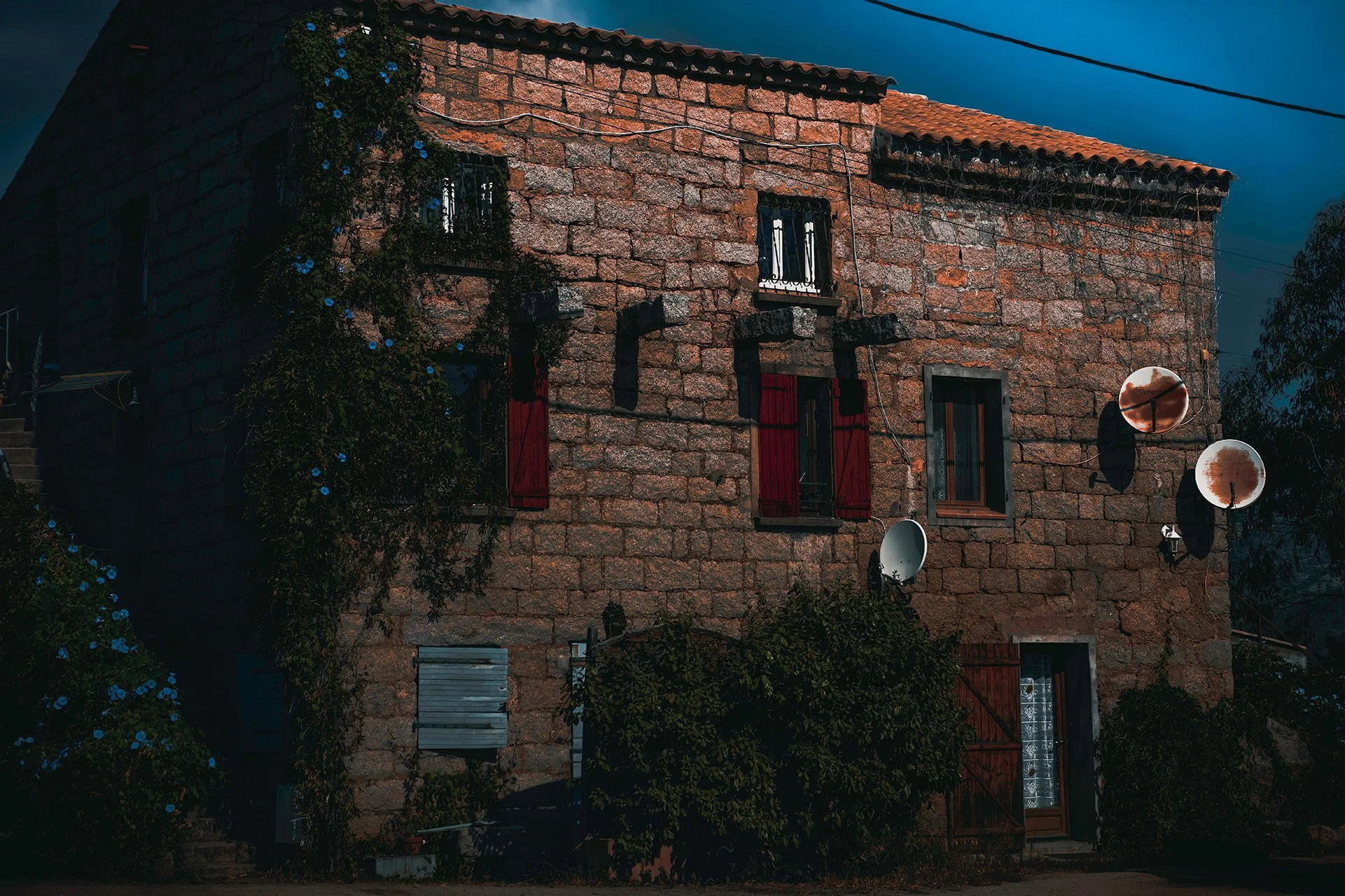 A brick house at night with closed red shutters on its windows, multiple satellite dishes attached to the wall, dark sky, and some foliage around the base.