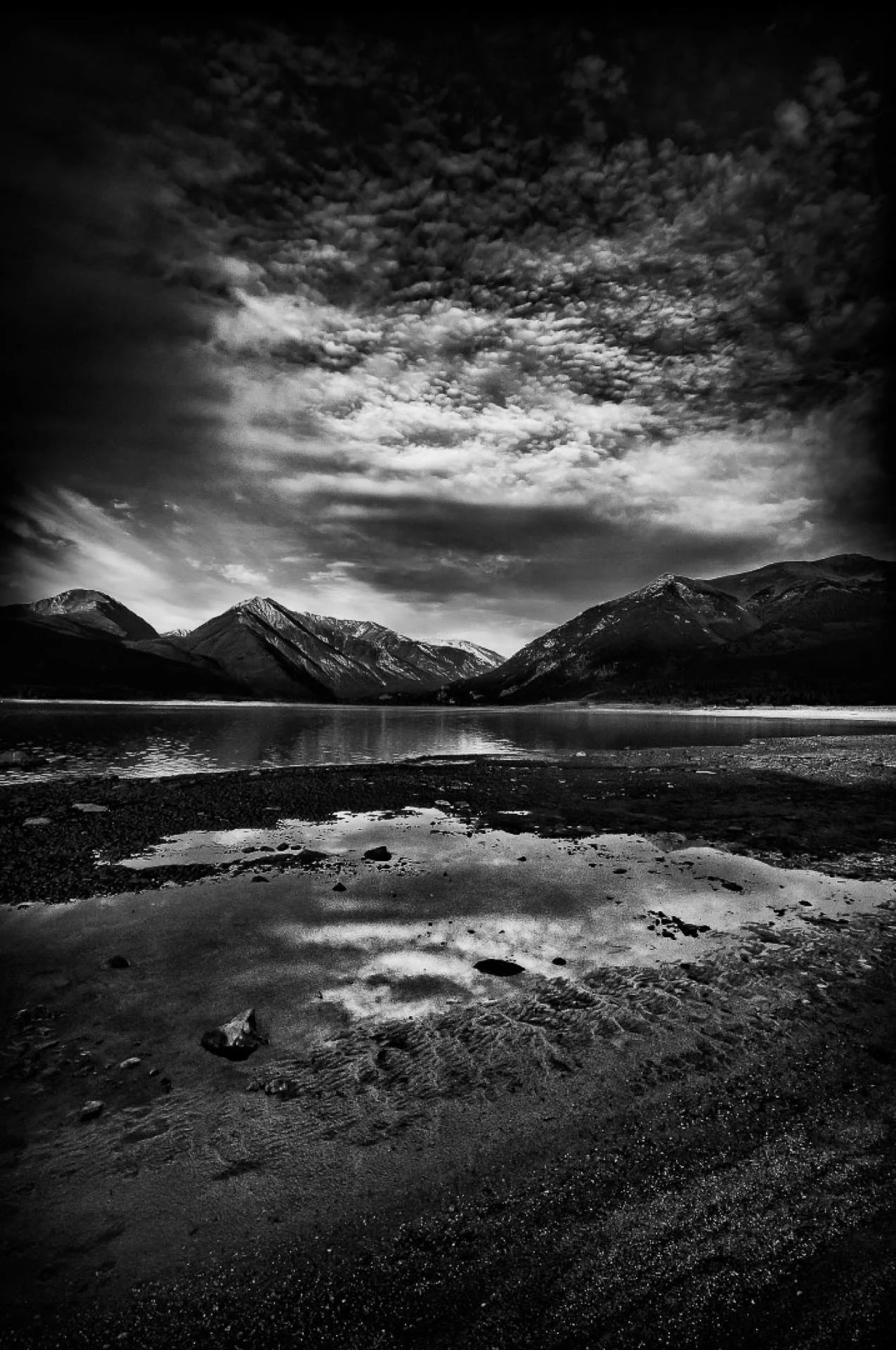 A black and white landscape photo of a mountain range reflecting in a body of water, with a cloudy sky overhead.
