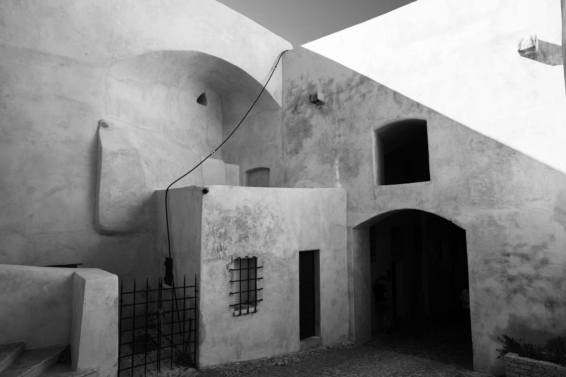 Black and white photo of a building with irregular, textured walls, arches, small barred windows, and a black door, featuring a simple railing and some plants outside.