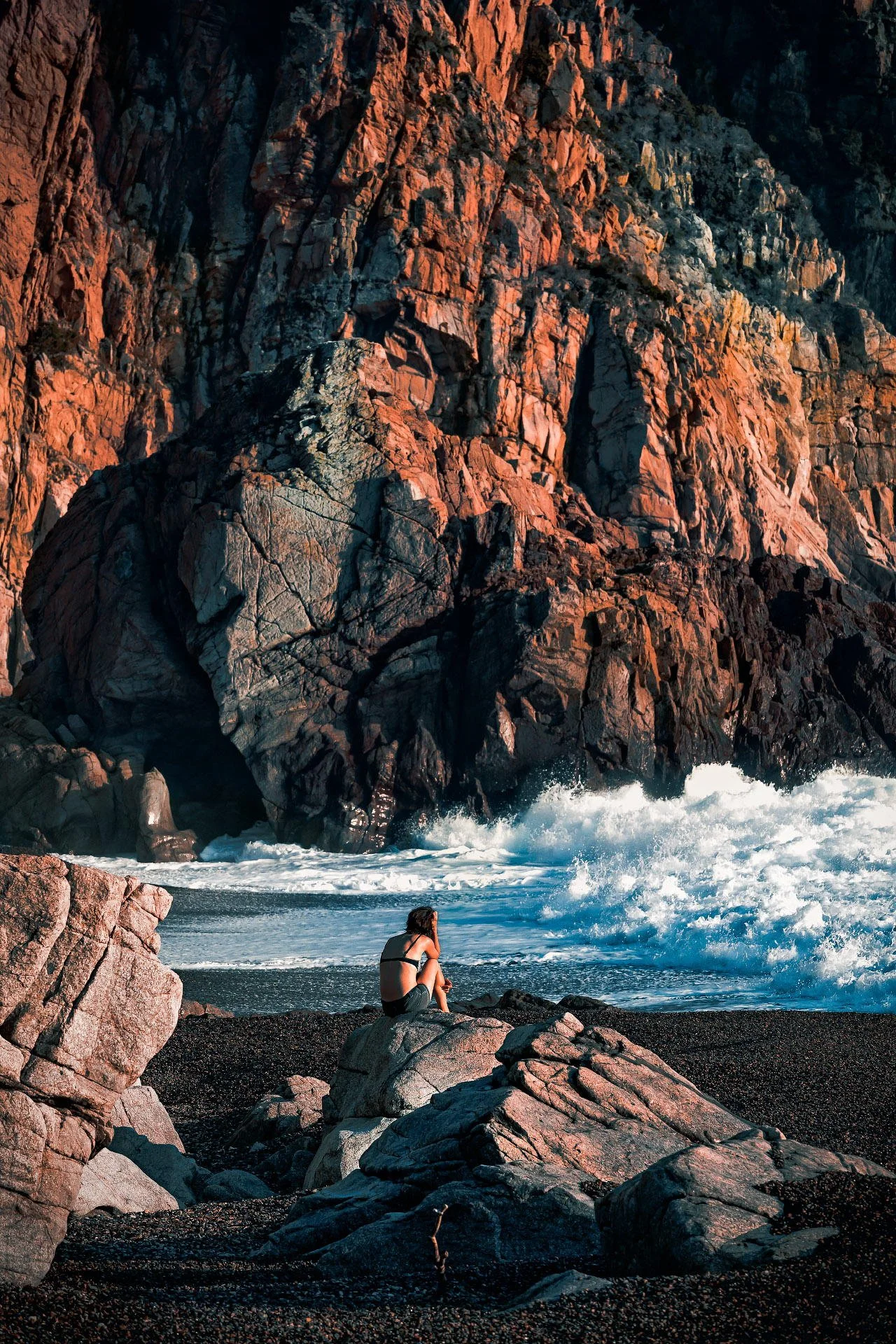 A woman sitting on rocks at the beach, facing the ocean waves with large orange and black rocks and cliffs in the background.